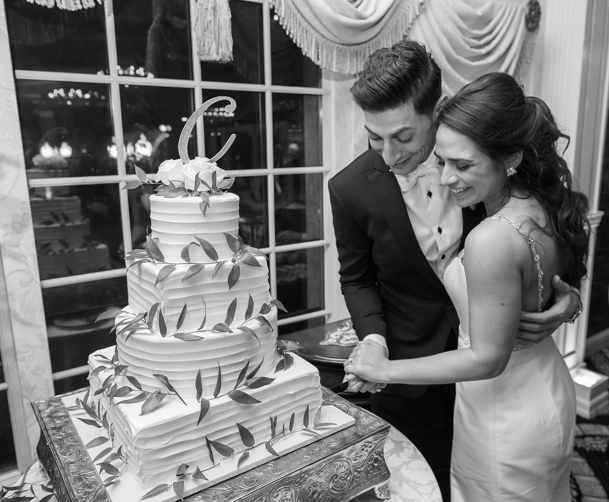 A couple is cutting a wedding cake together at a reception. The cake is decorated with leaves and flowers, positioned on a decorative table. The background features a window with drapes.