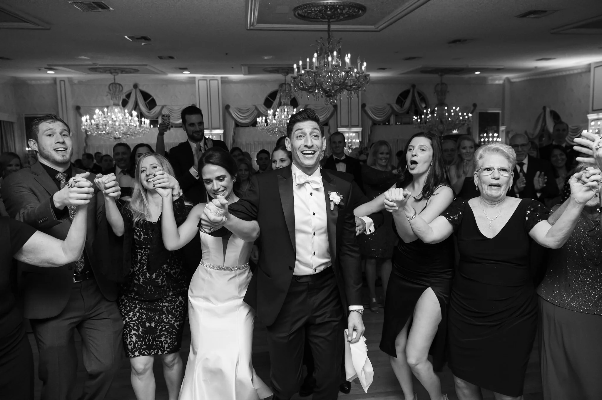 A group of people, including a bride and groom, holding hands and smiling at a celebration or wedding reception in an elegant ballroom with chandeliers.