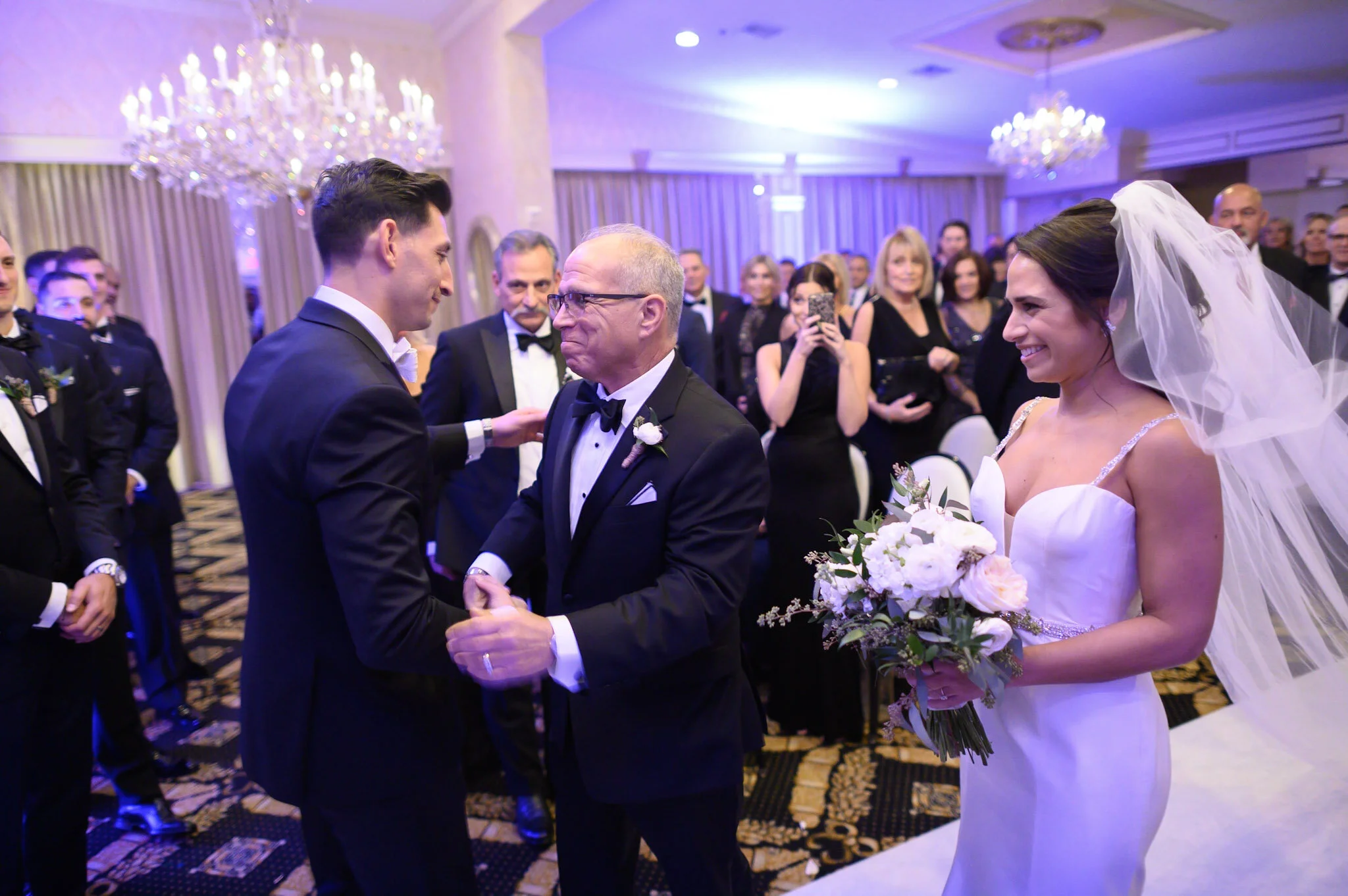 A wedding ceremony with a bride and groom exchanging vows. The bride is wearing a white wedding dress with a veil and holding a bouquet of white flowers. The groom is dressed in a black tuxedo. An officiant stands between them. Guests in formal attir