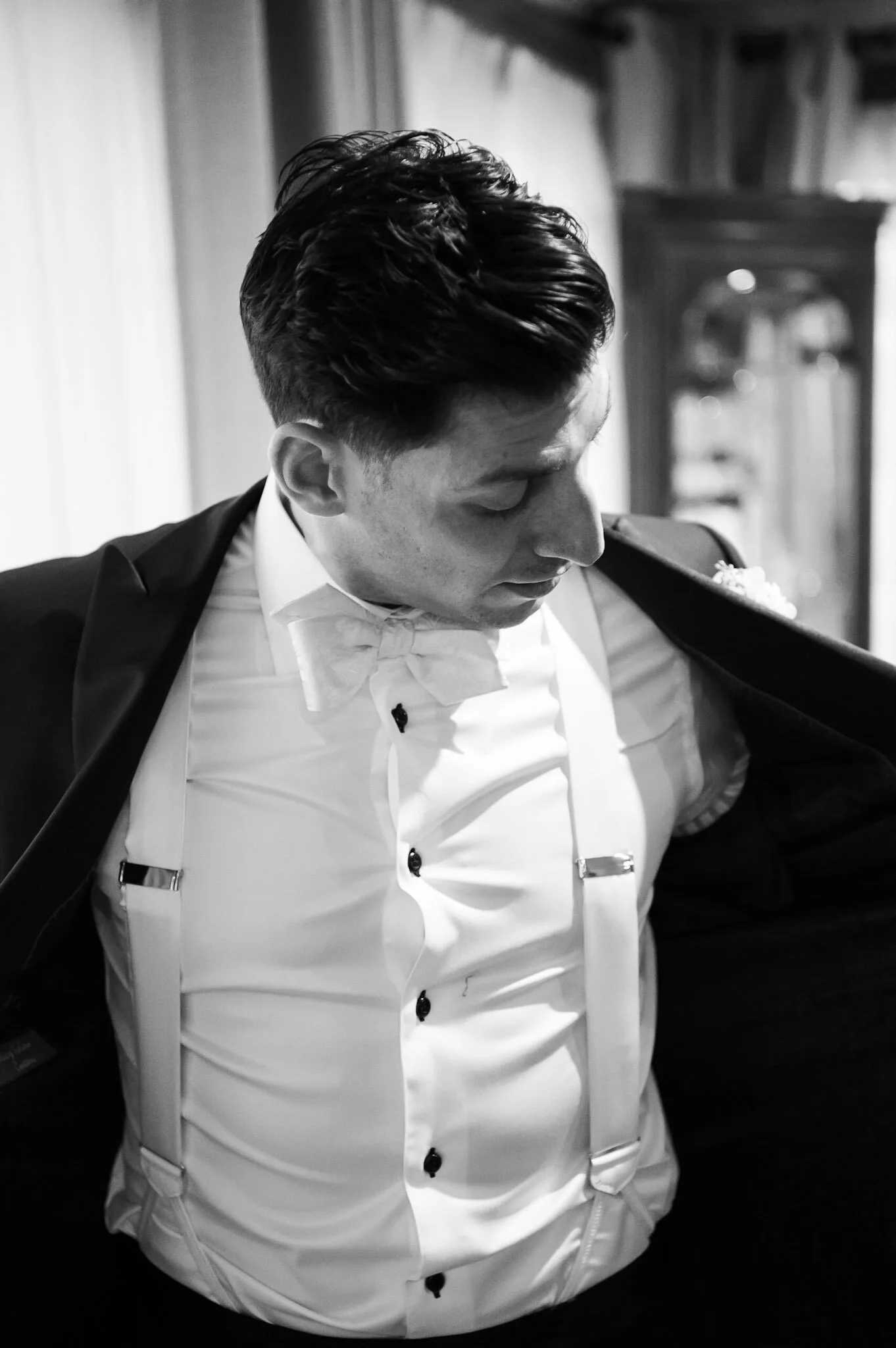A black and white photo of a man dressed in formal attire, including a tuxedo, bow tie, white shirt, and suspenders. His hair is styled neatly, and he is looking down, with a blurred background featuring a mirror or cabinet.