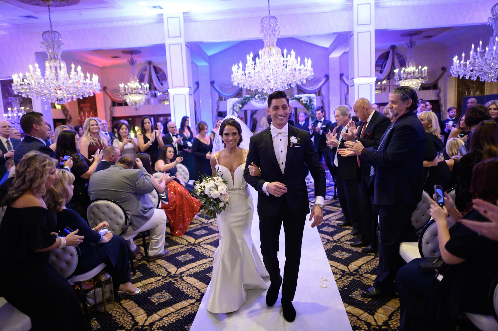 A bride and groom walking down the aisle at their wedding reception, surrounded by seated guests clapping and taking photos in an elegant venue with chandeliers and purple lighting.