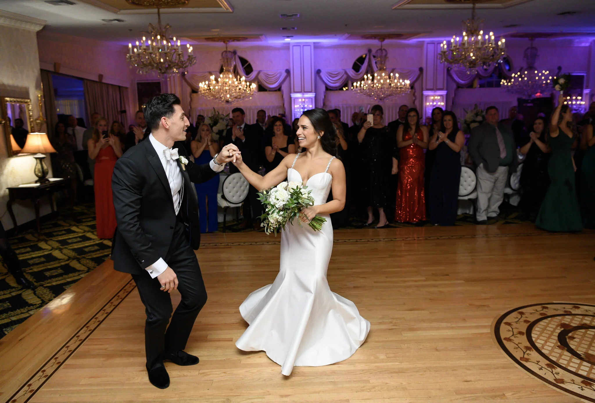 Bride and groom dancing at their wedding reception with guests clapping and smiling in the background.