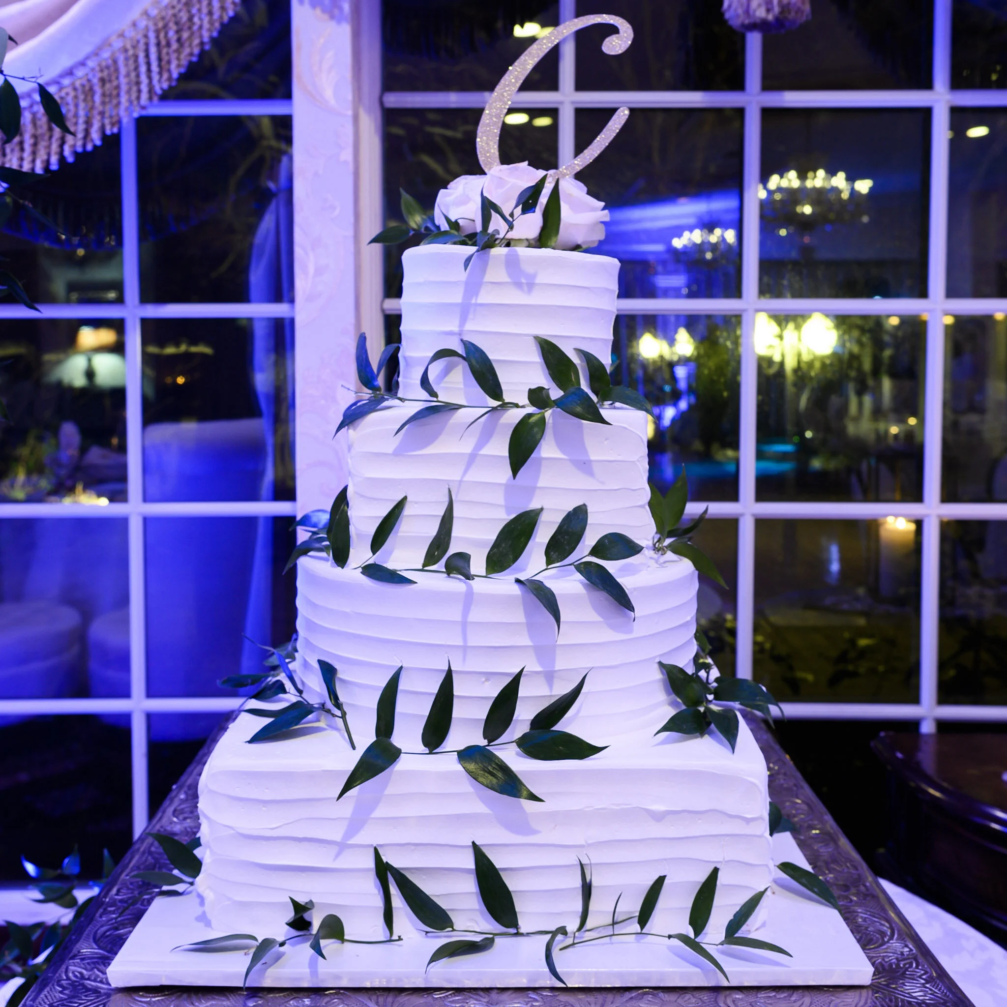 A decorative white tiered cake with green leaves placed on a silver tray, topped with a sparkling glittery number six and white flowers, illuminated with blue and yellow lights in the background.