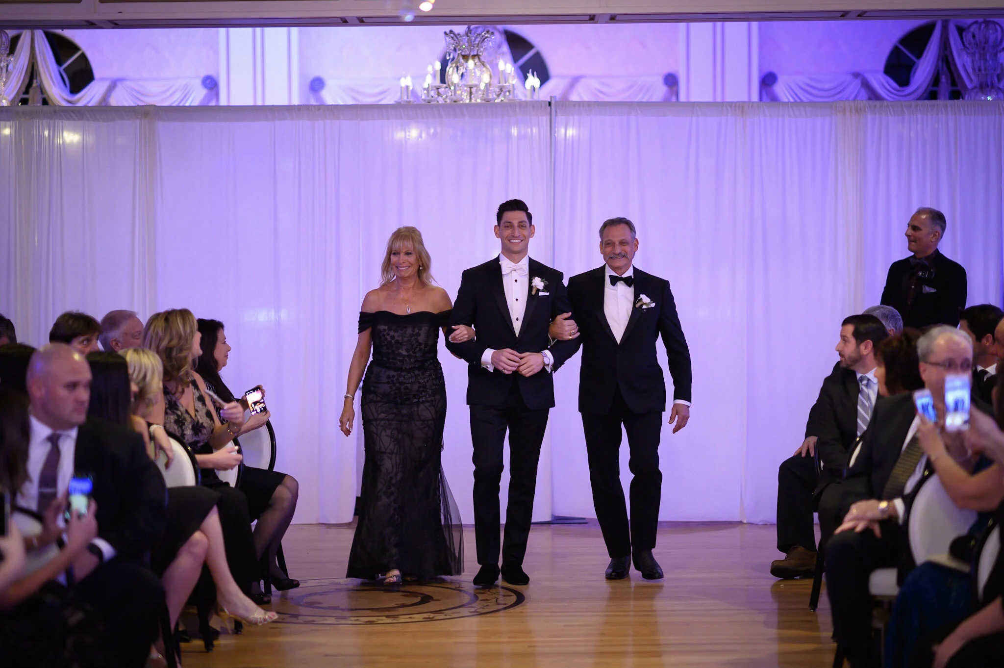 A bride and groom walking arm-in-arm down the aisle at their wedding ceremony, accompanied by an older woman, in a decorated indoor venue with guests seated on both sides.