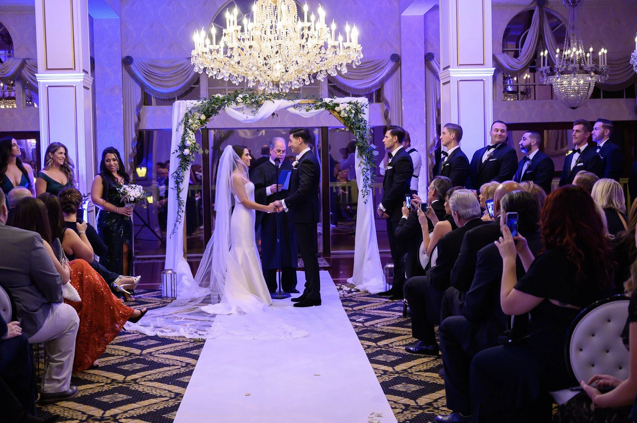A wedding ceremony inside a grand hall with chandeliers, white floral arch, bride and groom holding hands, officiant in black robes, bridesmaids in green dresses, groomsmen in tuxedos, guests seated and taking photos.