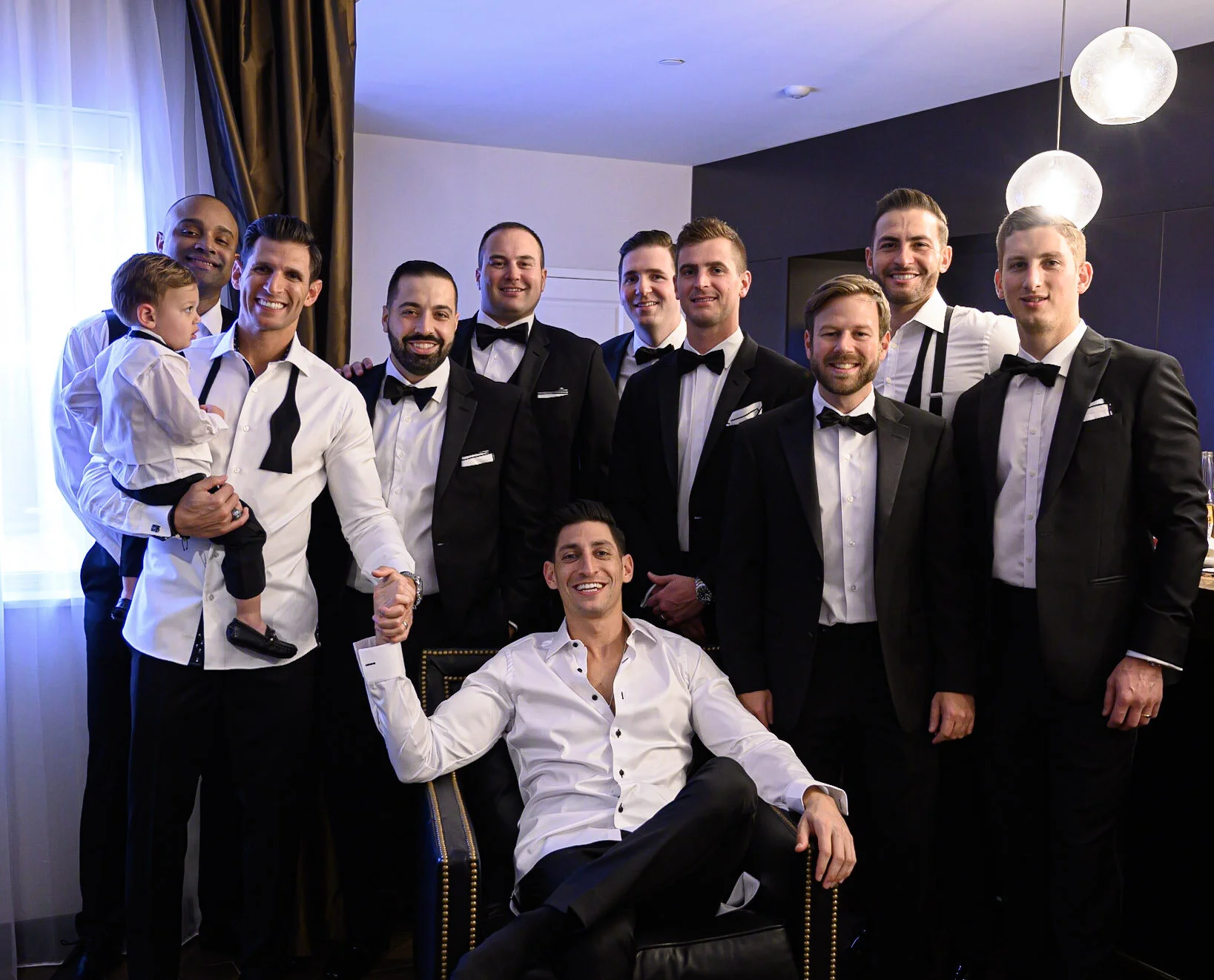 A group of men, most wearing tuxedos with bow ties, and one man in a white dress shirt, posing indoors in front of a dark wall with modern lighting. One man is seated in a chair, smiling with his arm resting on the chair's armrest. Another man holds 