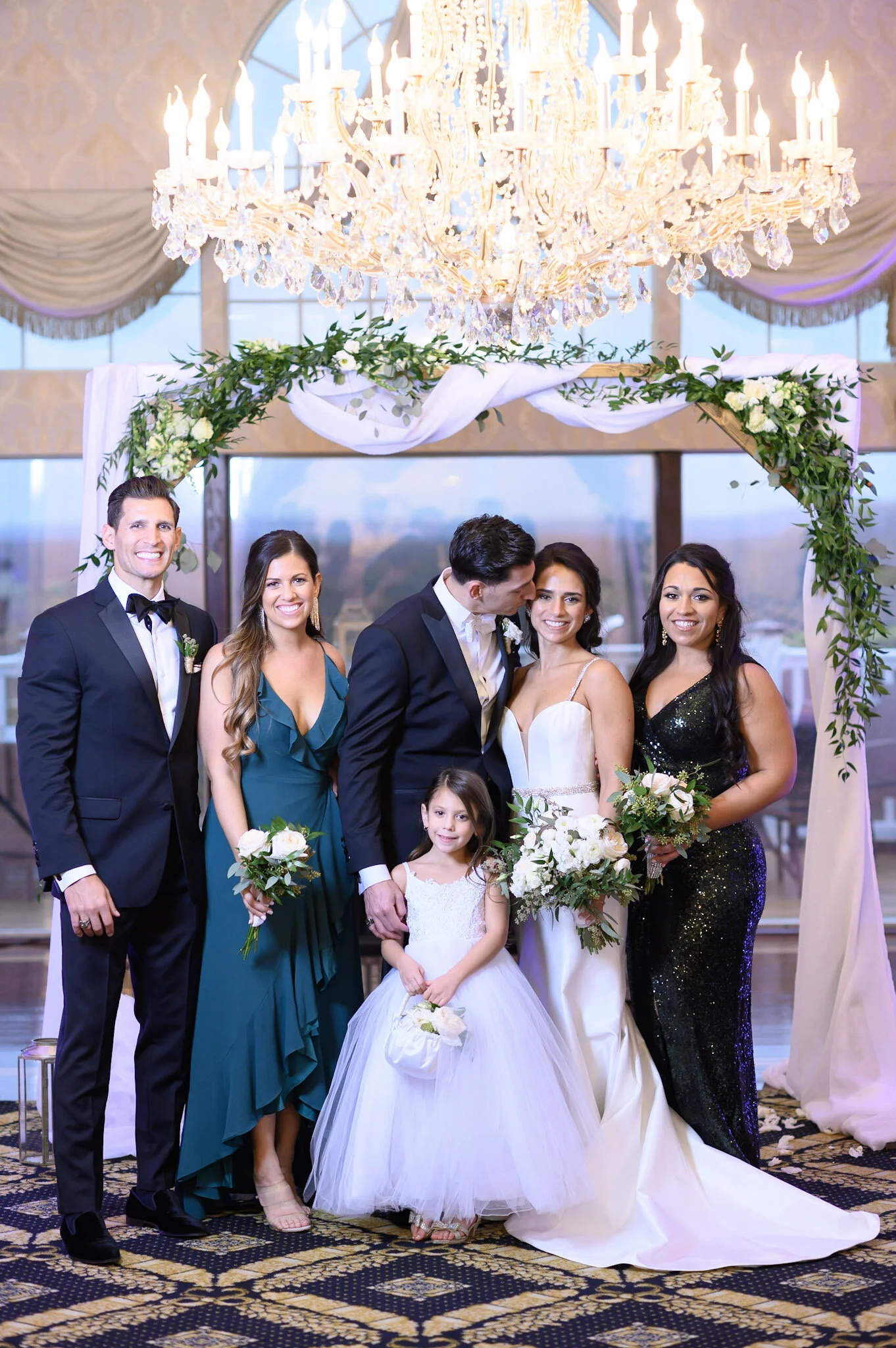 A group of people at a wedding, including a bride in a white gown holding a bouquet, a groom in a black tuxedo, and several guests in evening attire, standing under a decorated arch with flowers and greenery, in front of a large window and a grand ch