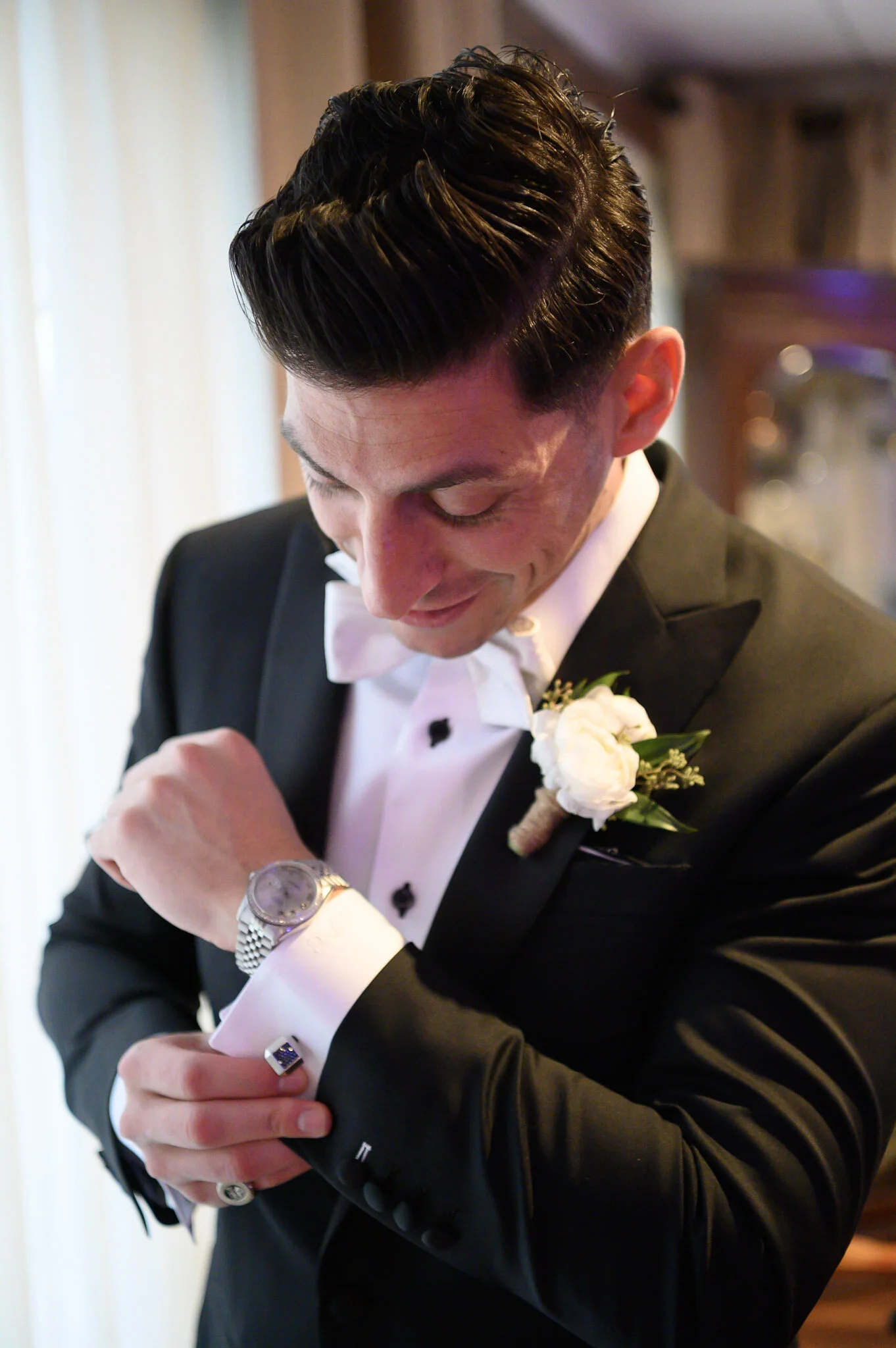A young man in a tuxedo with a white bow tie looks at his wristwatch while adjusting his sleeve, with a white flower boutonniere on his lapel and a patterned cuff link visible on his shirt.