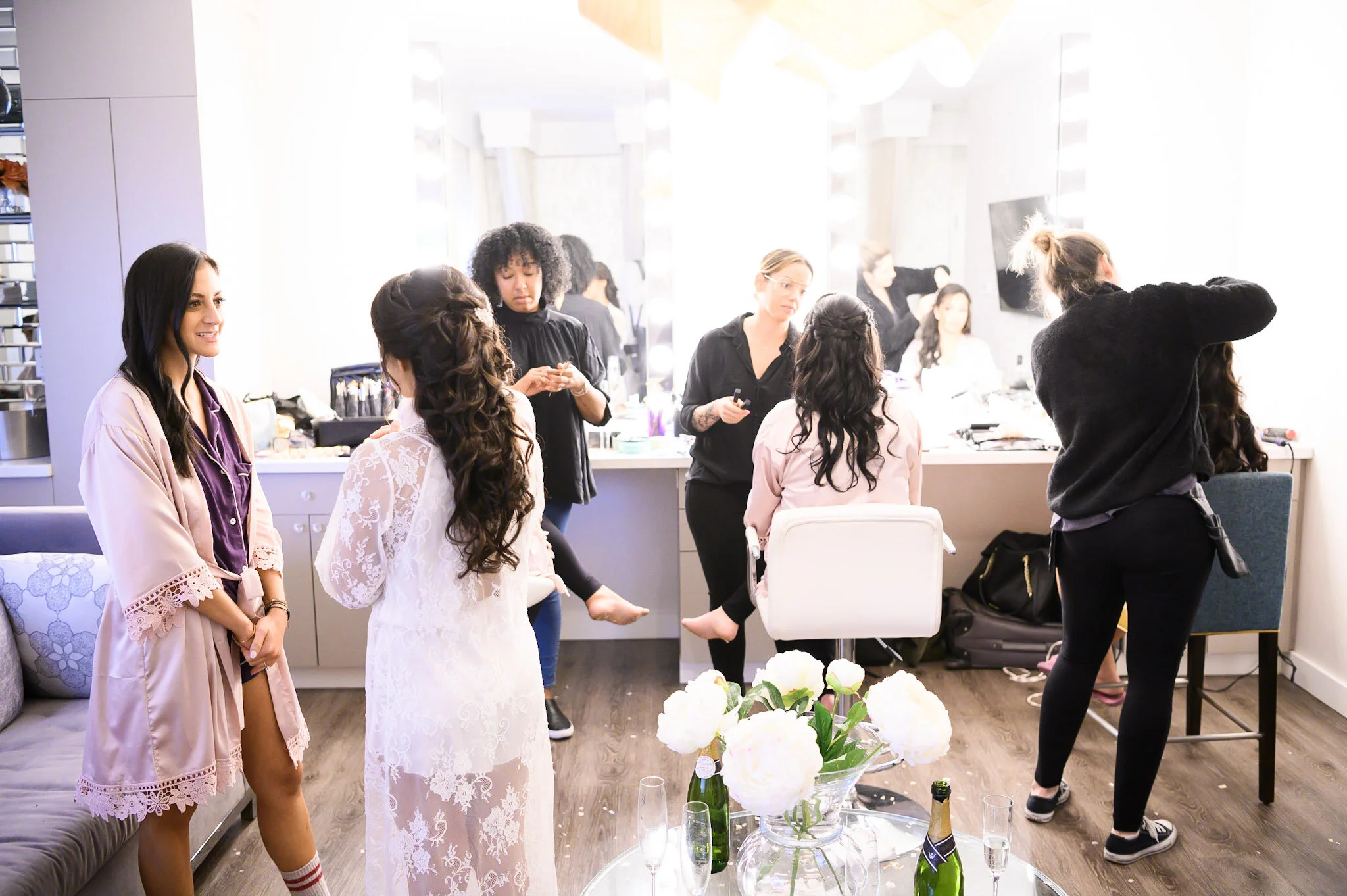 Women preparing for a wedding or special event in a well-lit dressing room with large mirrors, makeup and hair styling tools, and a table with flowers and champagne bottles.