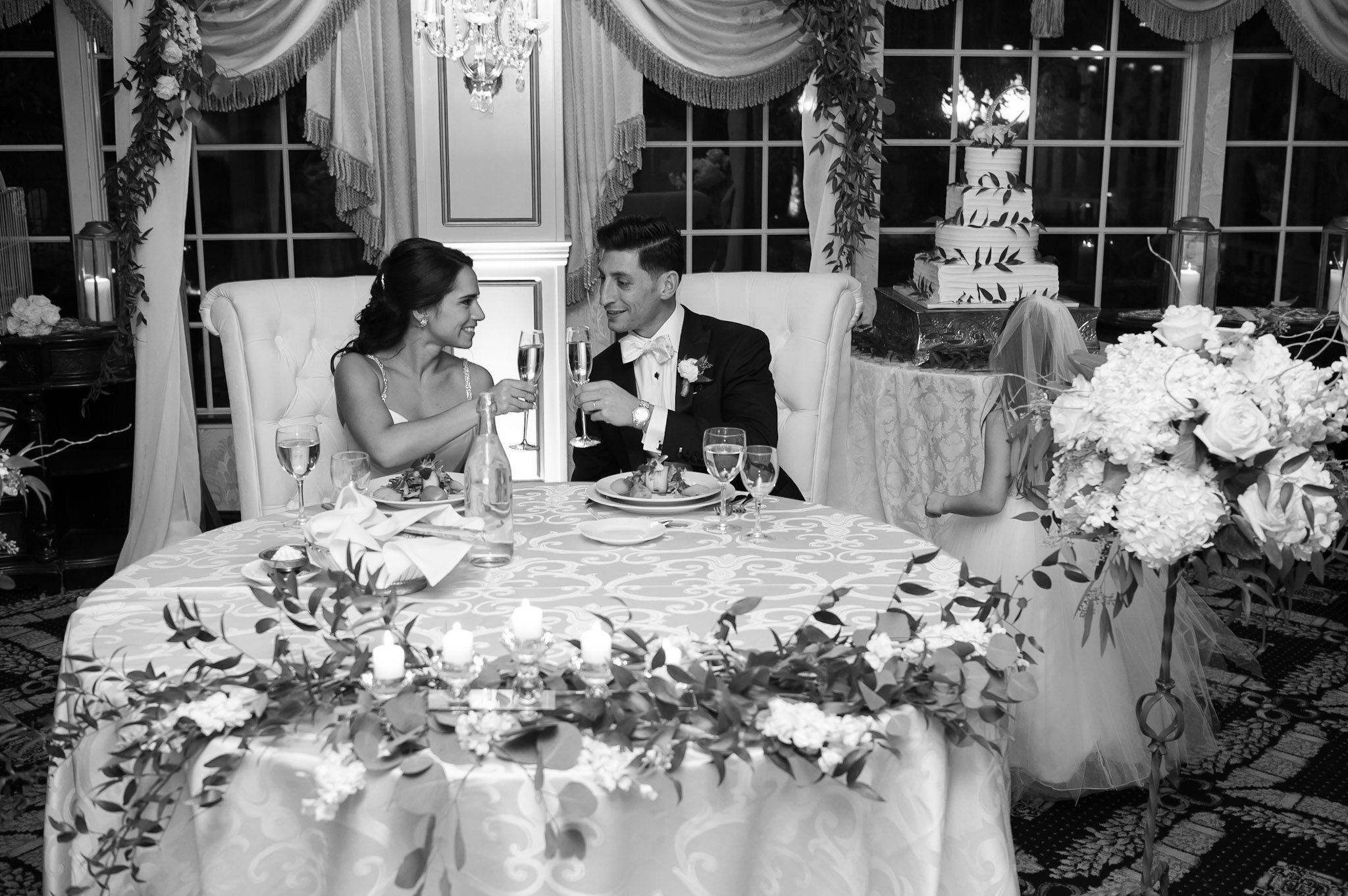 Black and white photo of a bride and groom holding glasses of champagne at a wedding reception, sitting in front of a decorative backdrop with bouquet flowers and wedding cake.
