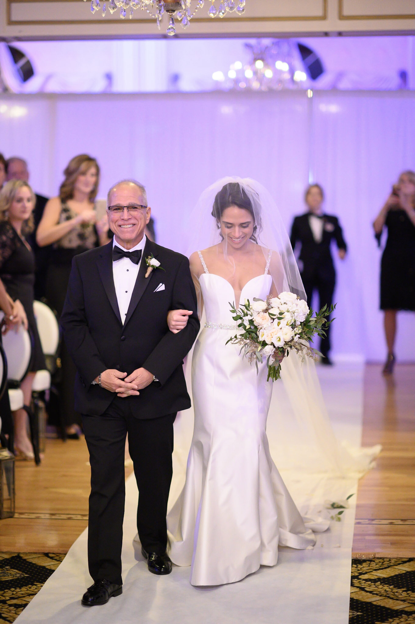 A bride in a white wedding gown and veil walking down the aisle with an older man in a tuxedo, likely her father, during a wedding ceremony. Guests are visible in the background, with a chandelier hanging from the ceiling.