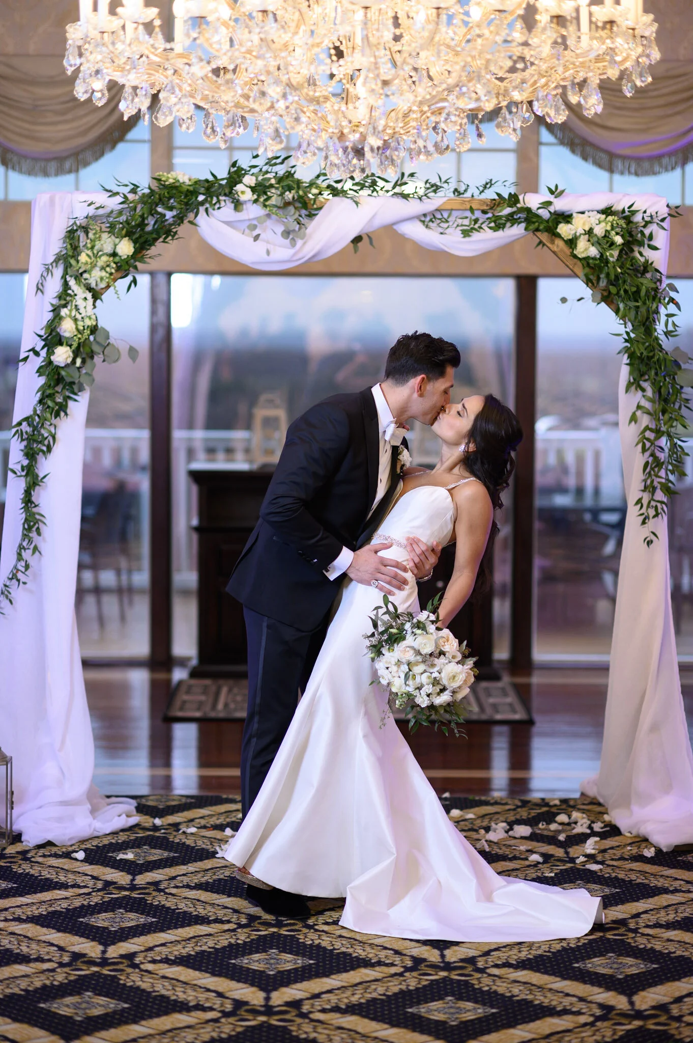 A bride and groom share a kiss at their wedding ceremony beneath a floral arch with a chandelier overhead.