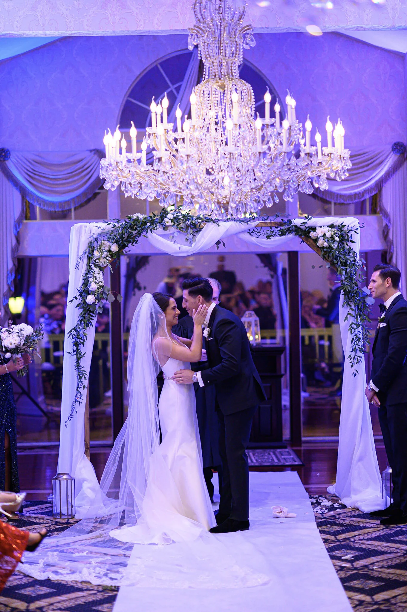 A bride and groom exchanging vows under a decorated arch with white drapery and green foliage, illuminated by a large crystal chandelier, during their wedding ceremony.