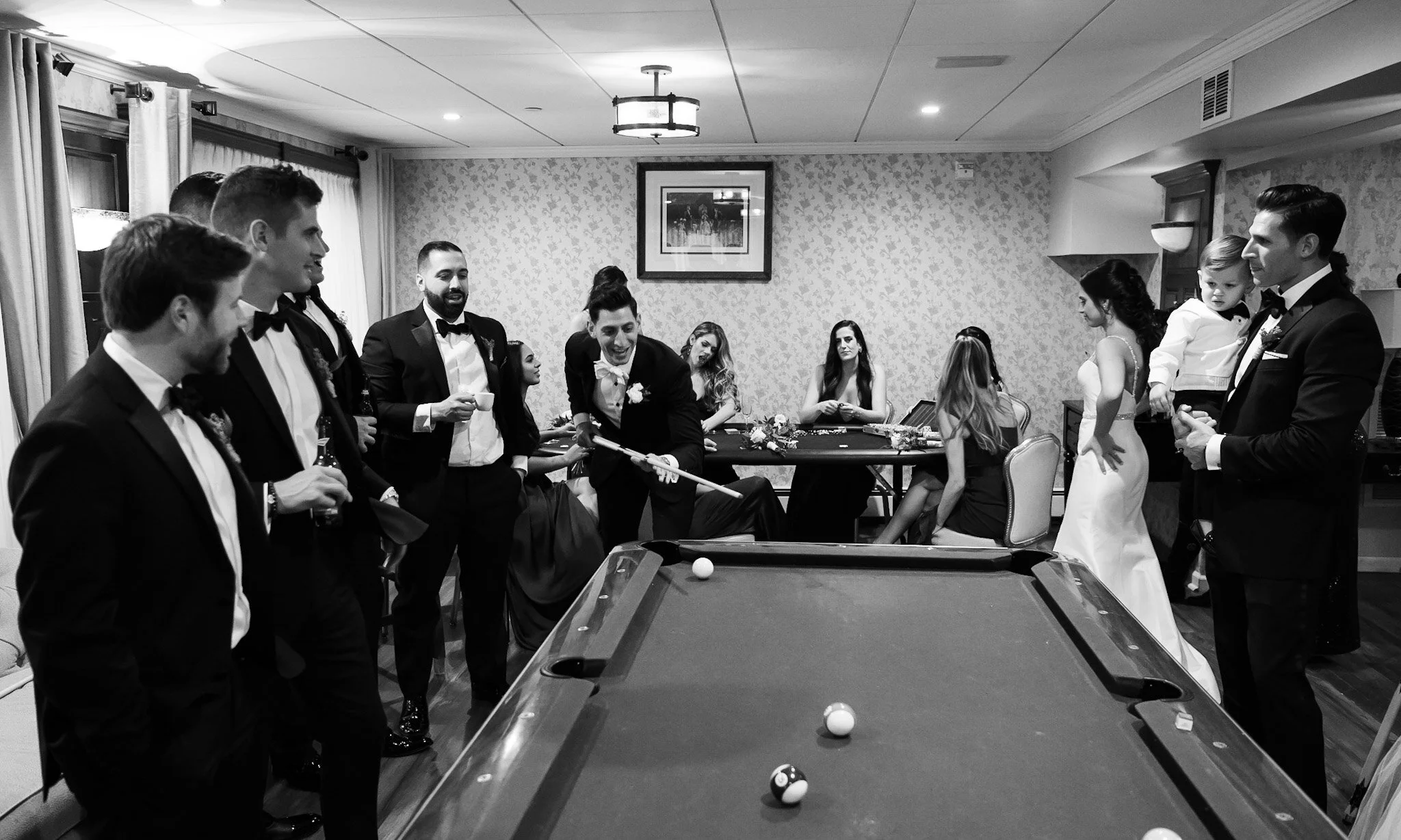 A group of well-dressed people at a formal event playing pool in a decorated room. Some are standing around the pool table, smiling and watching the game, while a man is about to take a shot.