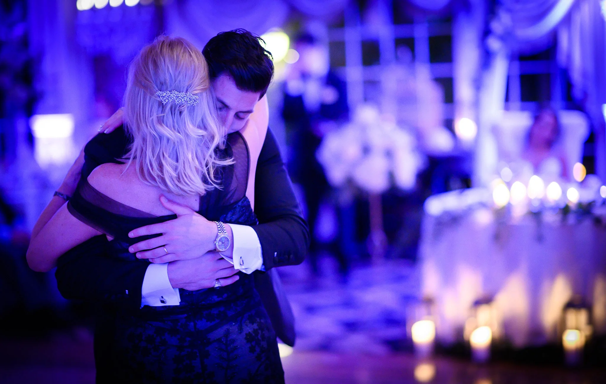 A couple dancing closely at a formal event with dim lighting and purple-blue hues, candles in the background.