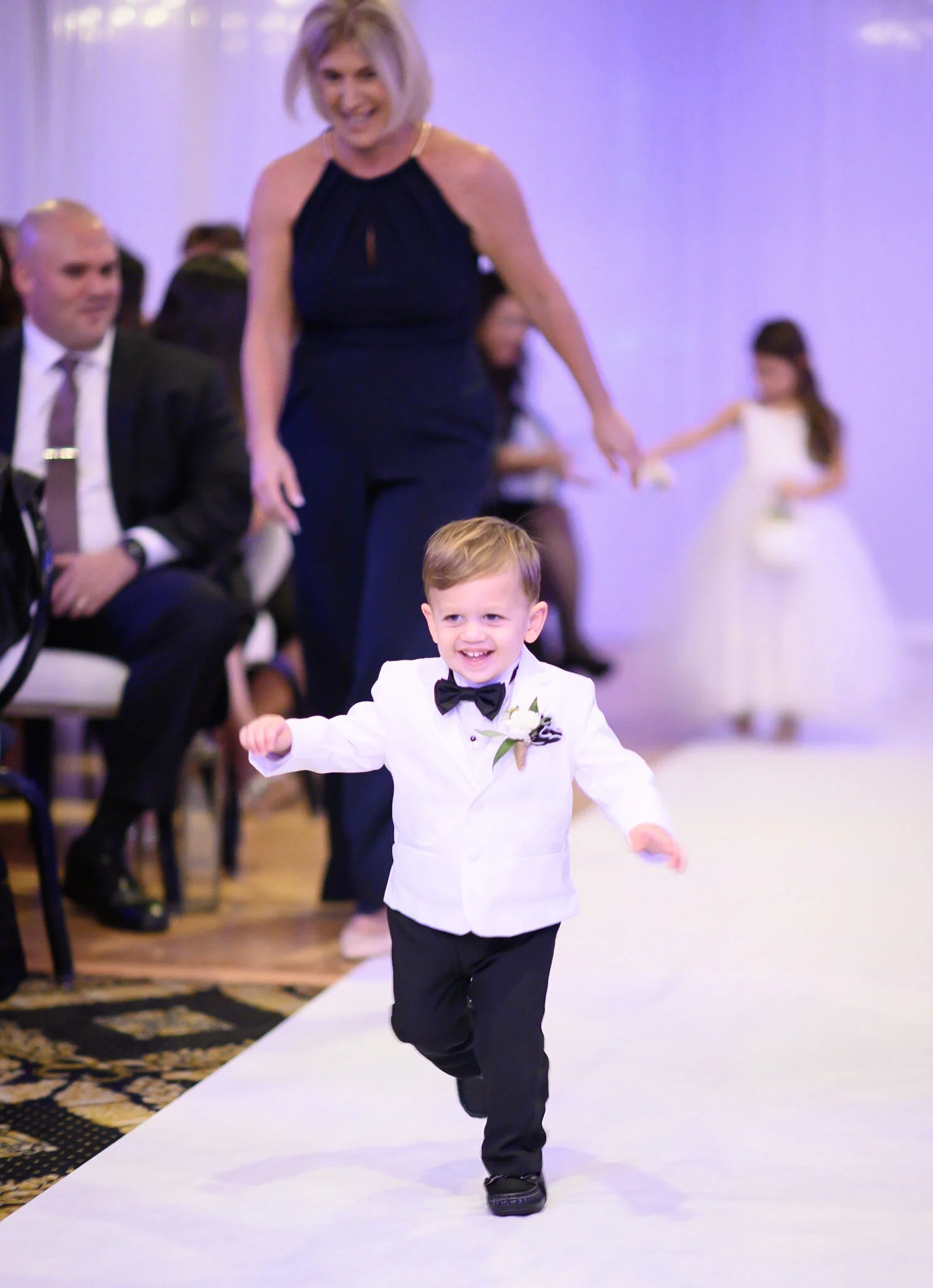 A smiling young boy in a white tuxedo with a black bow tie walks down a white runway at a formal event, with guests seated on either side and a woman in a black dress in the background.