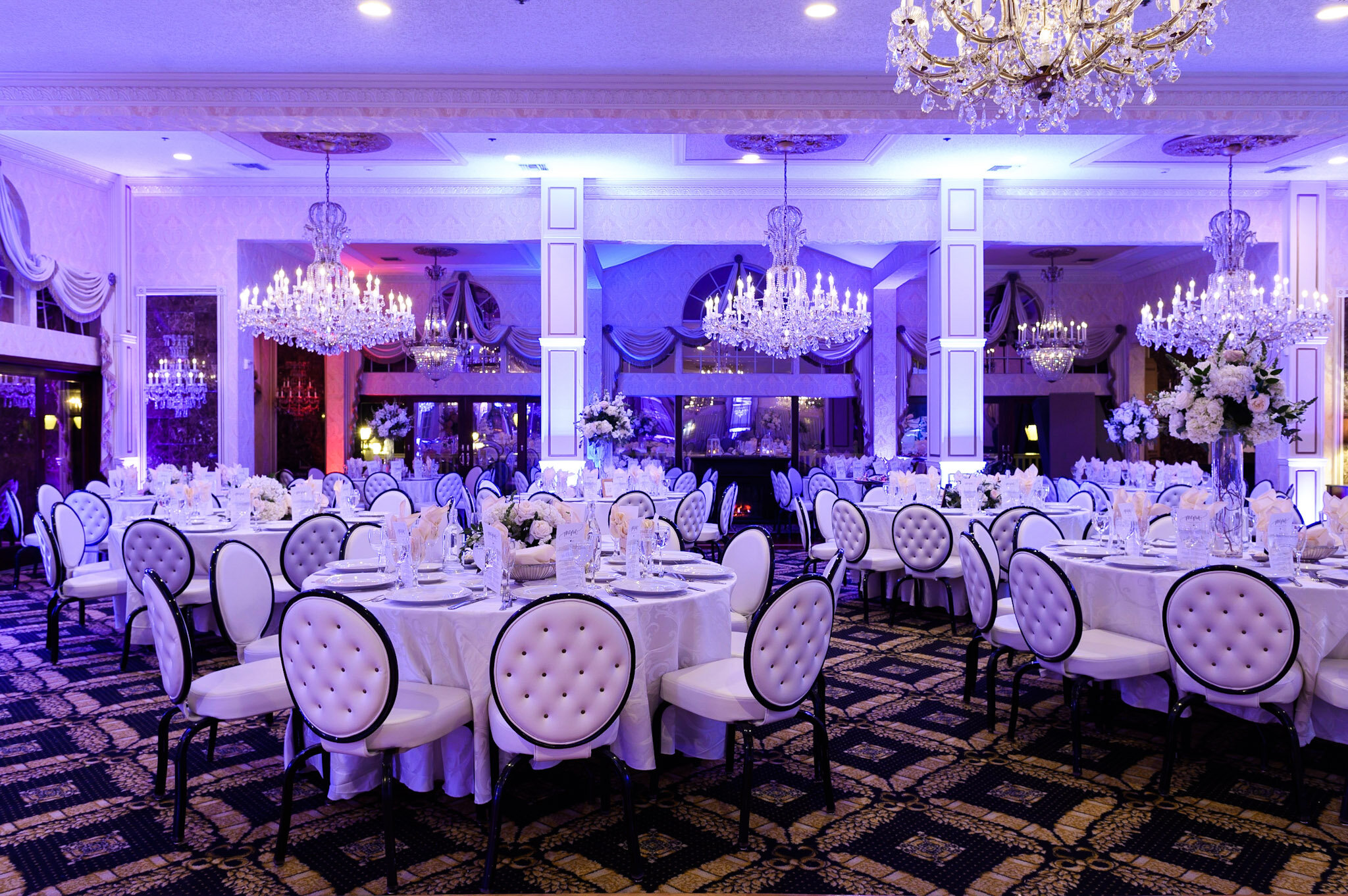 A banquet hall decorated with elegant chandeliers, large floral centerpieces, and ornate drapery. The room is set up with round tables covered with white cloths and white upholstered chairs, ready for a formal event.