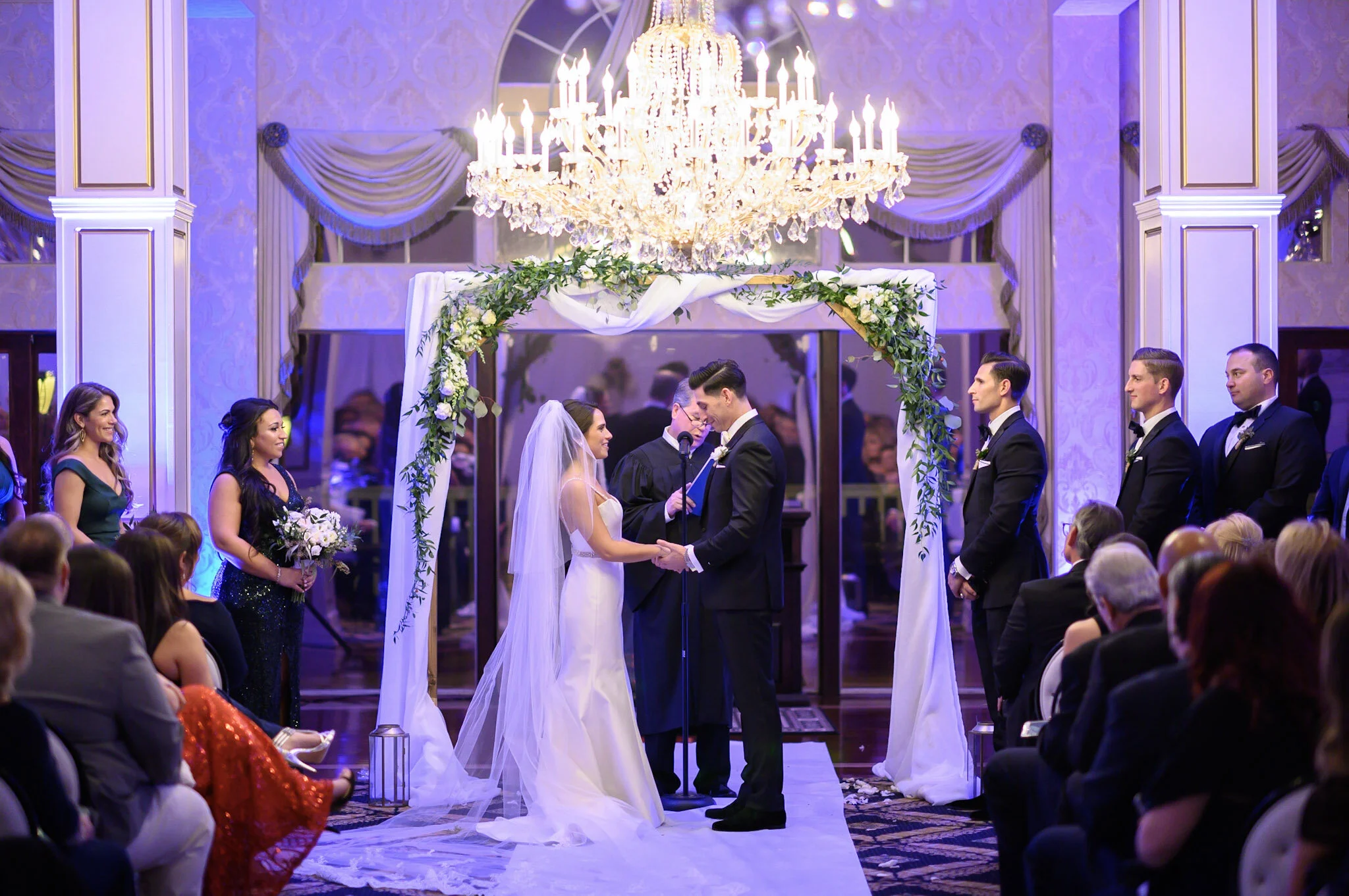 A wedding ceremony taking place indoors under a large crystal chandelier. The bride and groom are standing at the altar holding hands, with the officiant reading from a book. Bridesmaids and groomsmen are standing on either side, with guests seated a