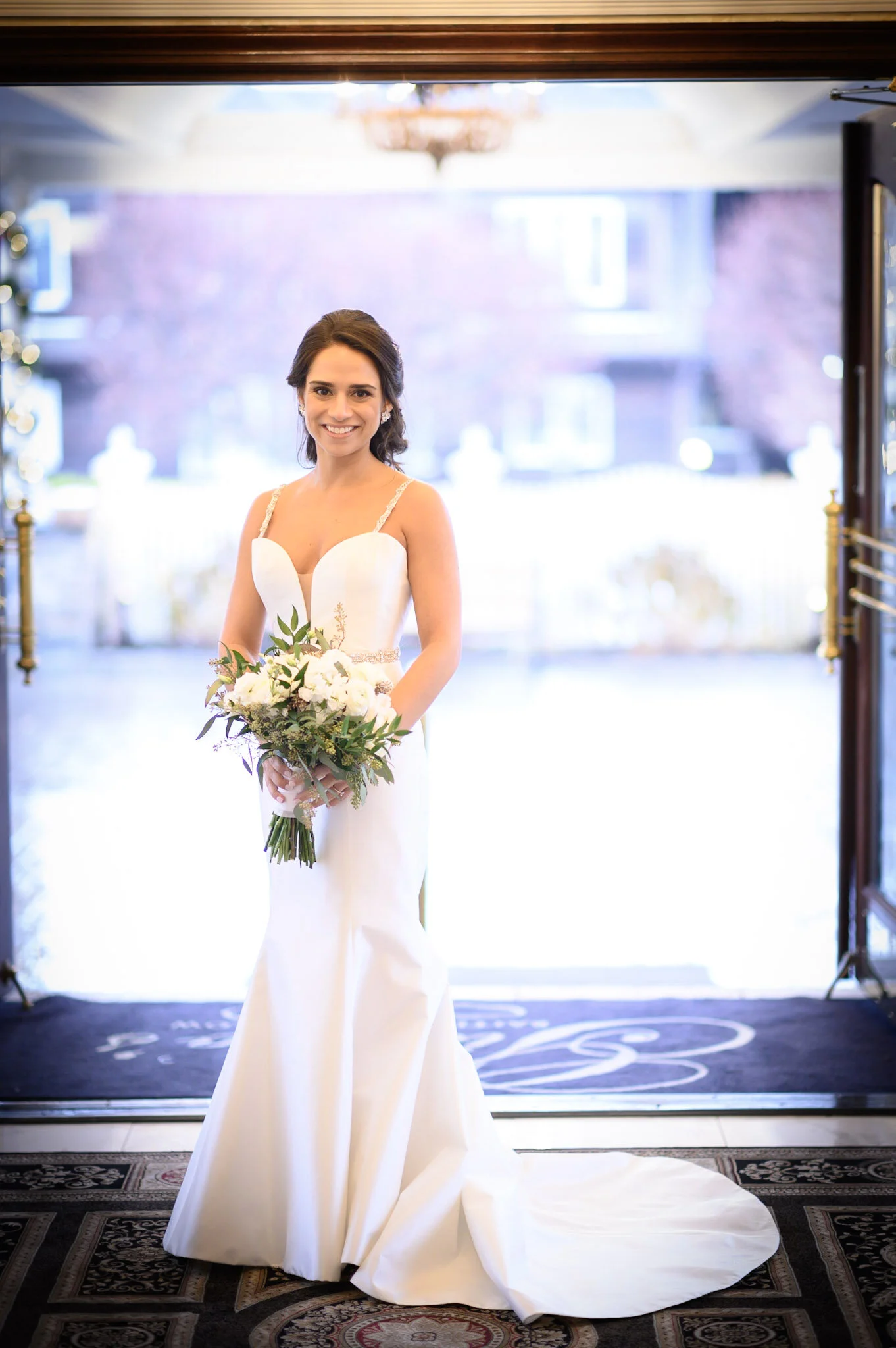 A smiling bride standing inside near the entrance, holding a bouquet of white flowers with greenery. She is wearing a white wedding dress with spaghetti straps, standing on a decorative rug with light coming from outside.