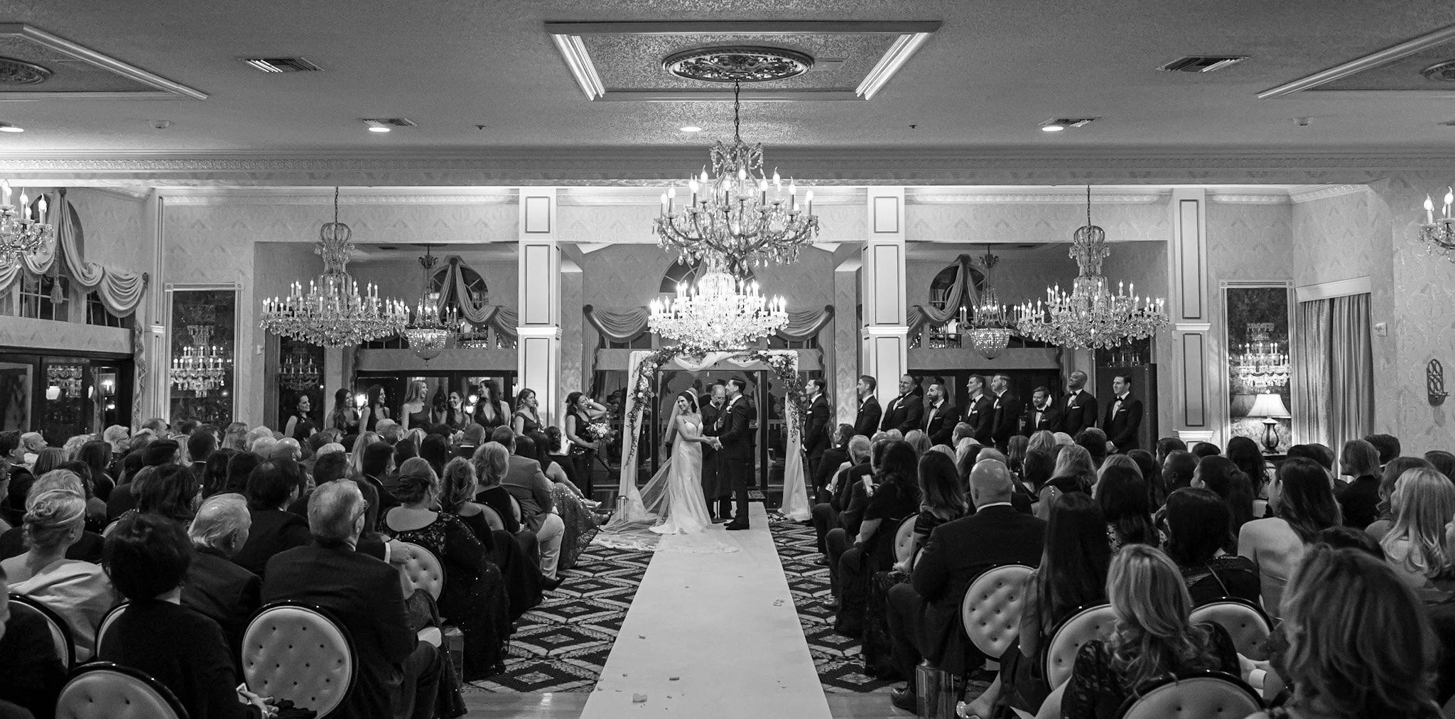 A black and white photo of a wedding ceremony with the bride and groom holding hands at the altar, surrounded by bridesmaids and groomsmen, in a grand banquet hall with chandeliers and ornate decorations, with guests seated facing the ceremony.