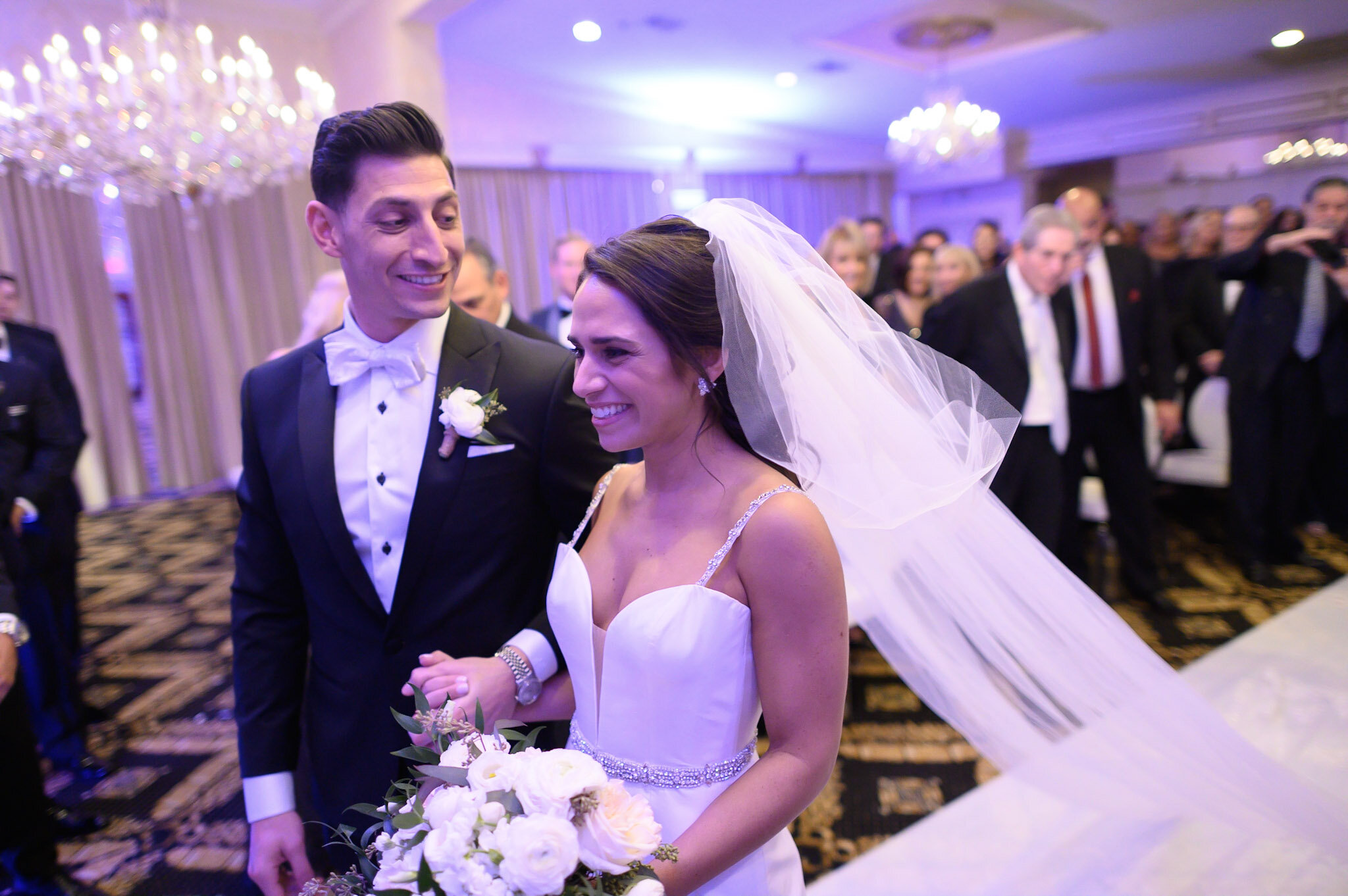 A bride and groom smiling at each other during their wedding reception, with guests dancing and socializing in the background.
