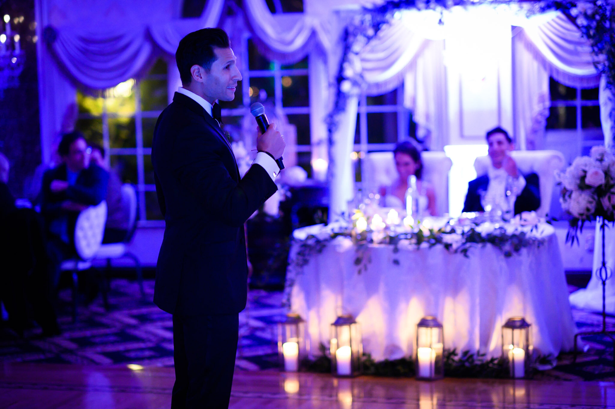 A man in a tuxedo holding a microphone giving a speech at a wedding reception in a decorated banquet hall. The background has dim purple lighting, large windows with draped curtains, and a long table with floral arrangements and candles.