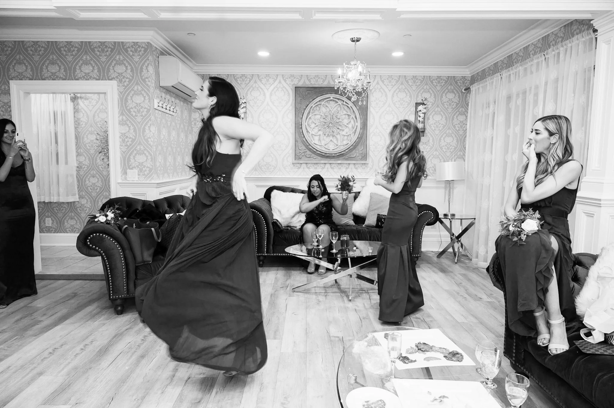 Women in formal dresses gathering in living room, some standing and talking, one sitting on a couch, a woman taking a photo, and a woman adjusting her dress, with glasses and drinks on a table.