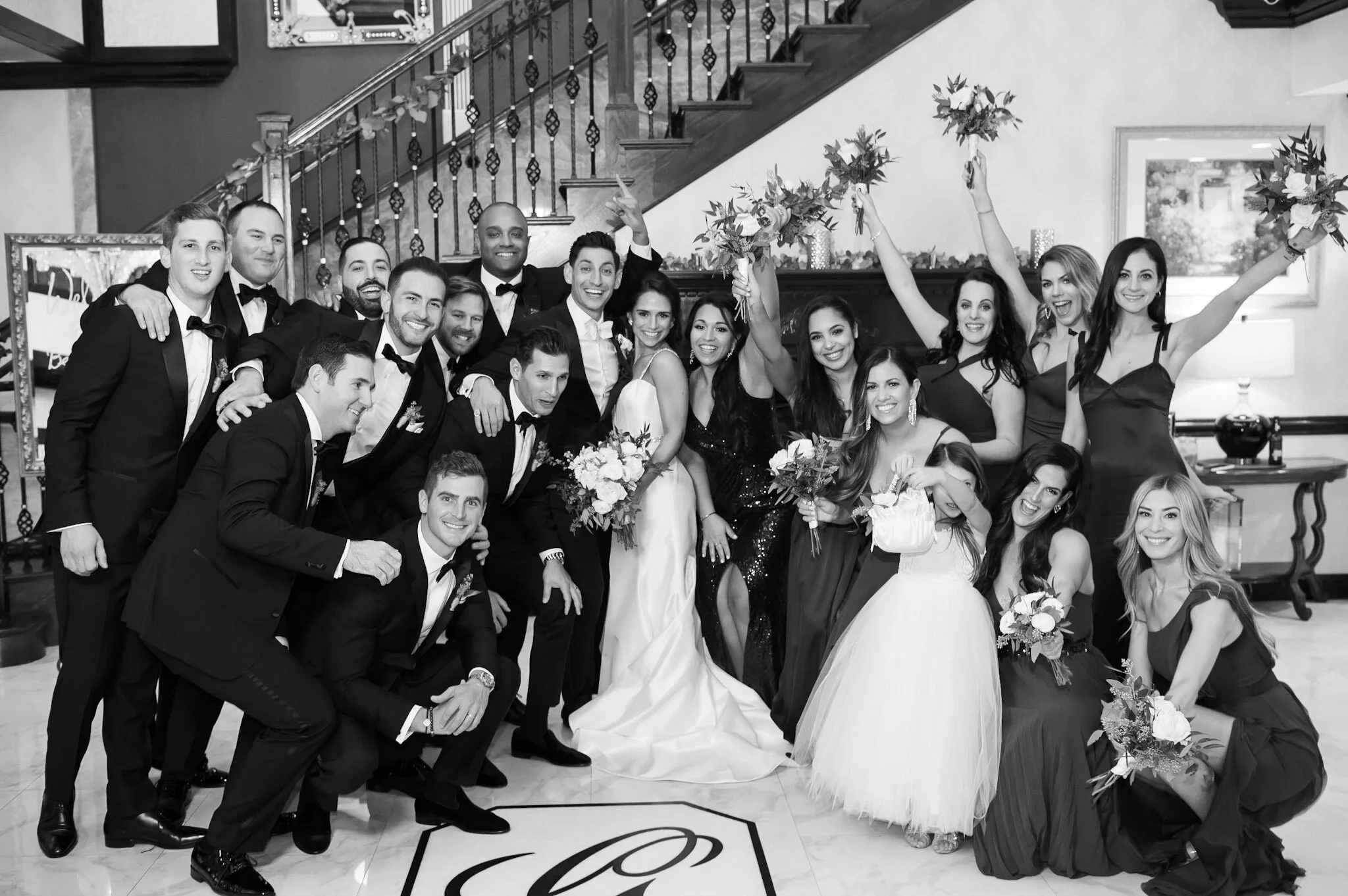 A group of people dressed in formal attire celebrating a wedding, with women holding bouquets and men in tuxedos, posing indoors on a staircase with a railing, decorated with flowers.