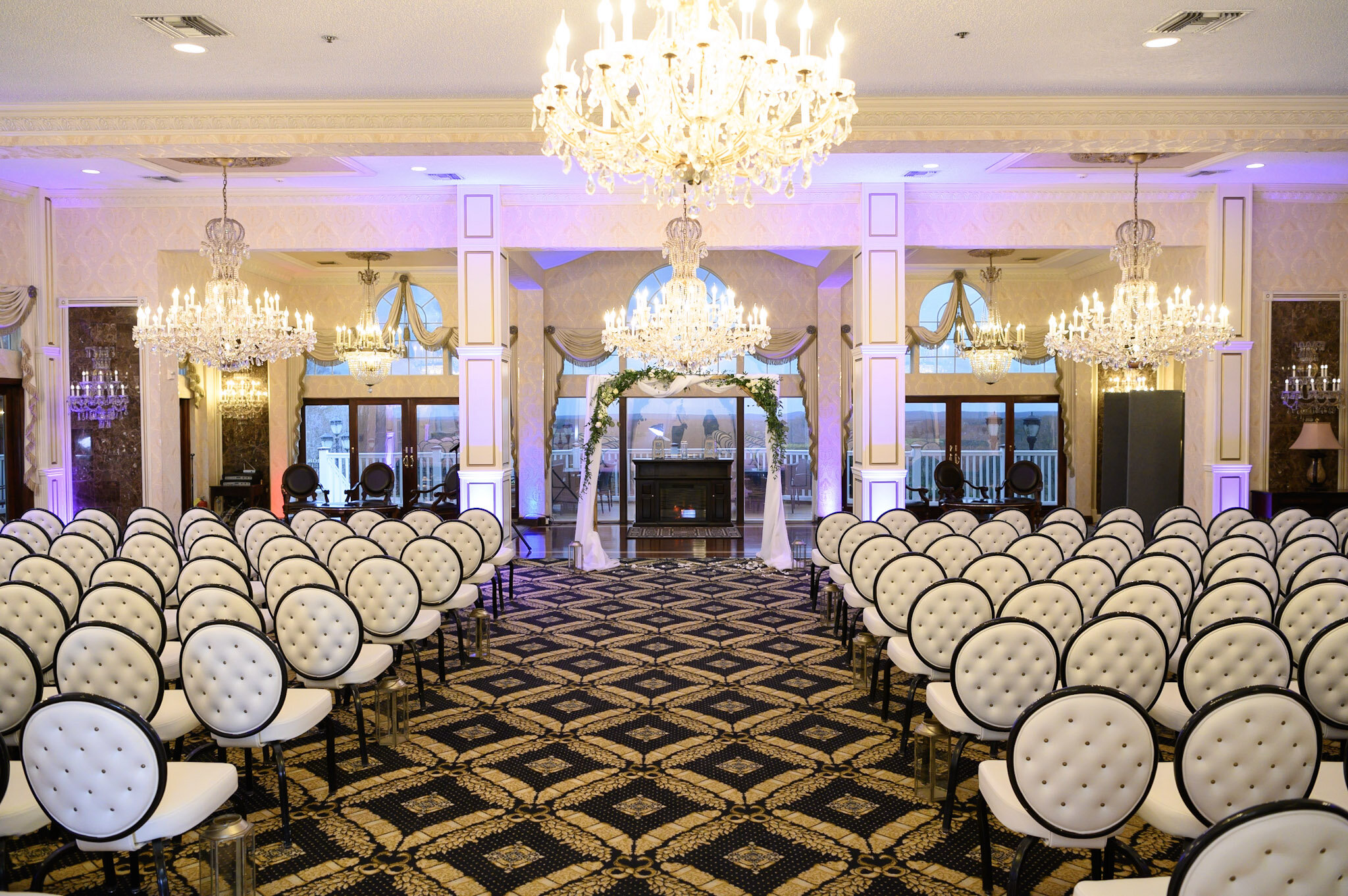 Elegant banquet hall decorated for a wedding with white chairs arranged in rows, crystal chandeliers, and a floral archway at the front.