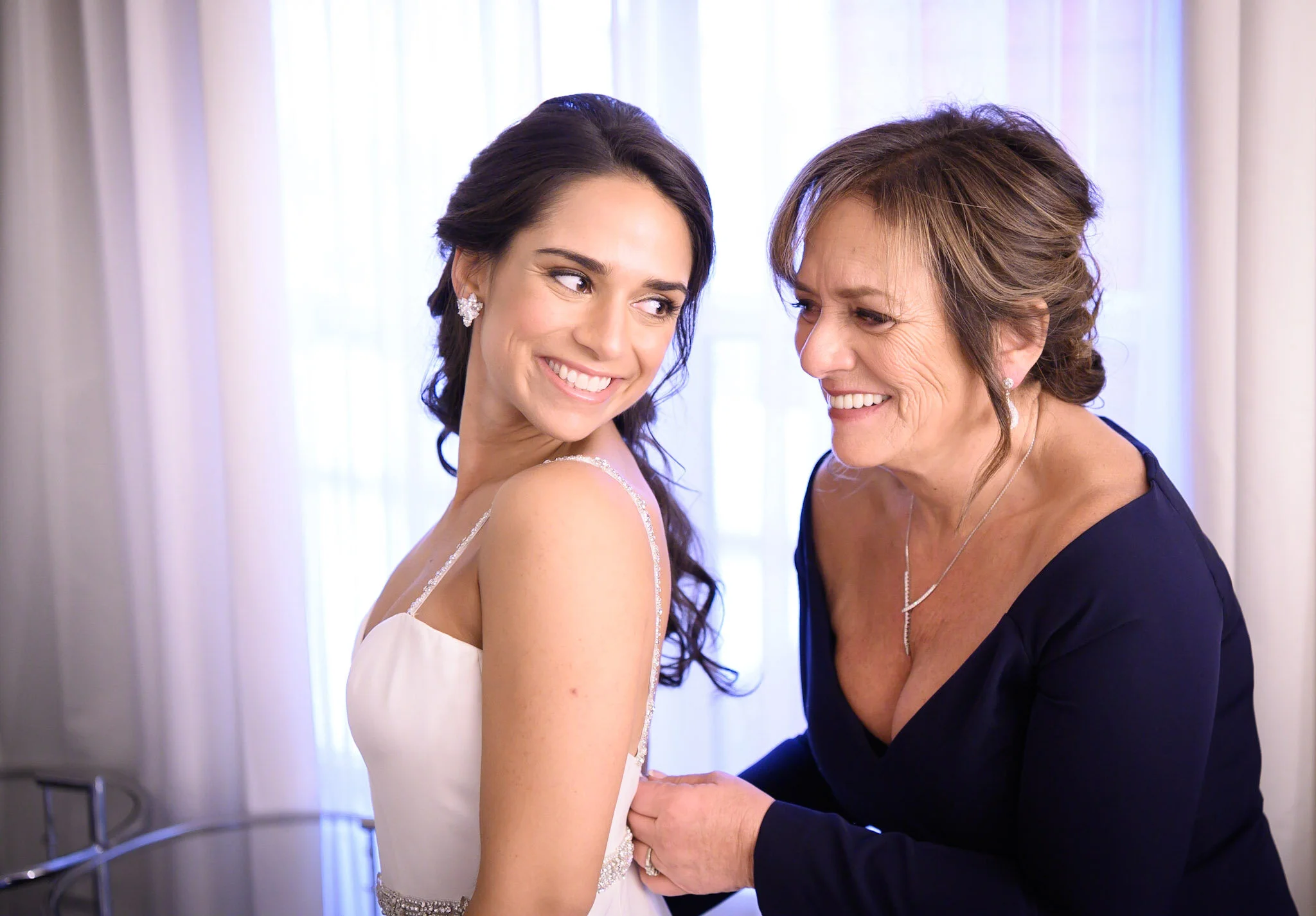A young woman in a white dress smiling at an older woman in a dark dress, both smiling and looking at each other.