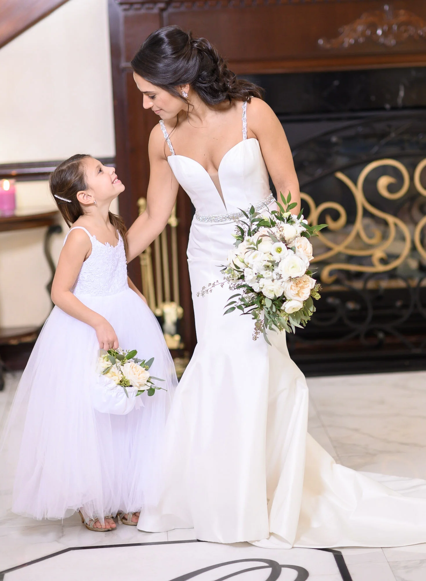 A bride in a white wedding dress holding a bouquet of flowers, looking at a young girl in a white dress holding a smaller bouquet, both standing indoors near a decorative black iron gate.