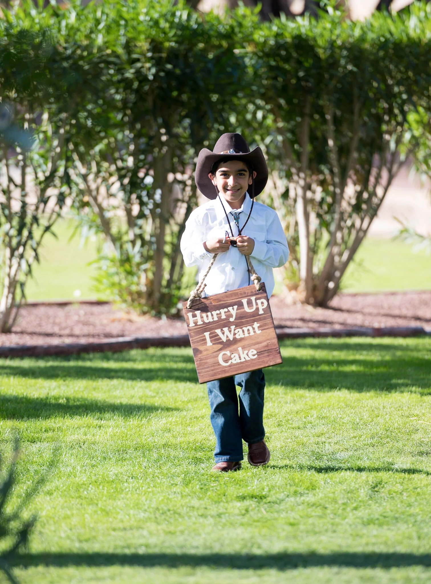 Young boy dressed as a cowboy, wearing a large cowboy hat, white shirt, blue jeans, and brown boots, walking on a sunny lawn with a wooden sign that says "Hurry Up I Want Cake" hanging around his neck, in front of a background of bushes and trees.