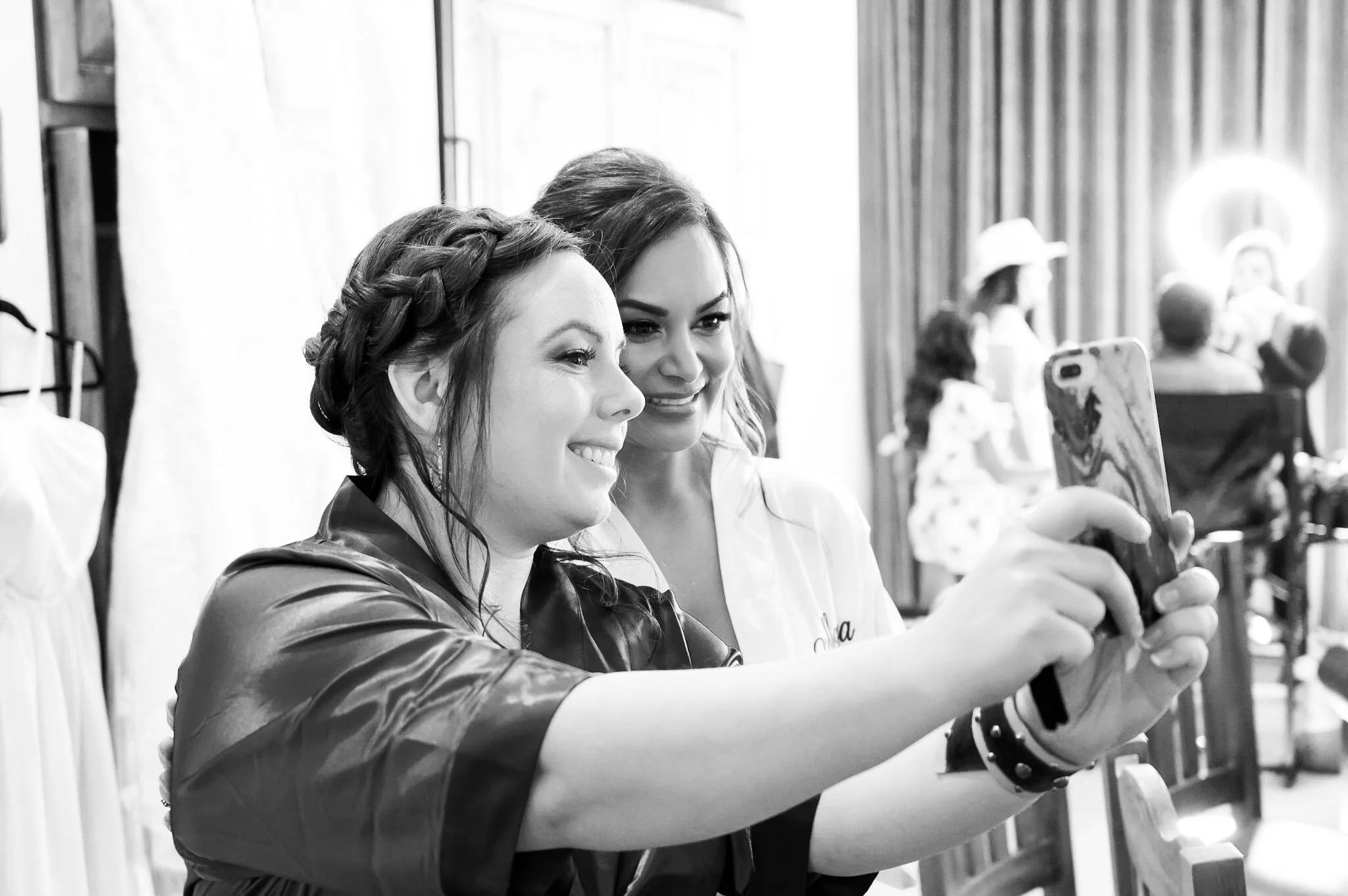 Two women are taking a selfie together in a well-lit room, smiling and looking at a smartphone. Other people can be seen in the background.