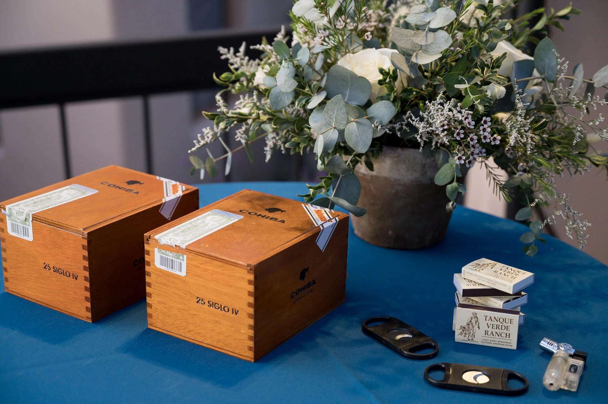 A blue table with three wooden cigar boxes, a potted plant with white flowers and green leaves, and various tools and supplies for tobacco rolling, including a small stack of cards, scissors, and a rolling device.
