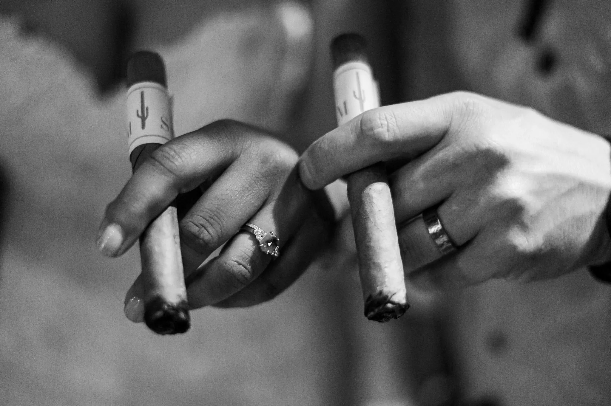 Close-up of two hands holding unlit cigarettes, with wedding rings visible on both hands in black and white.