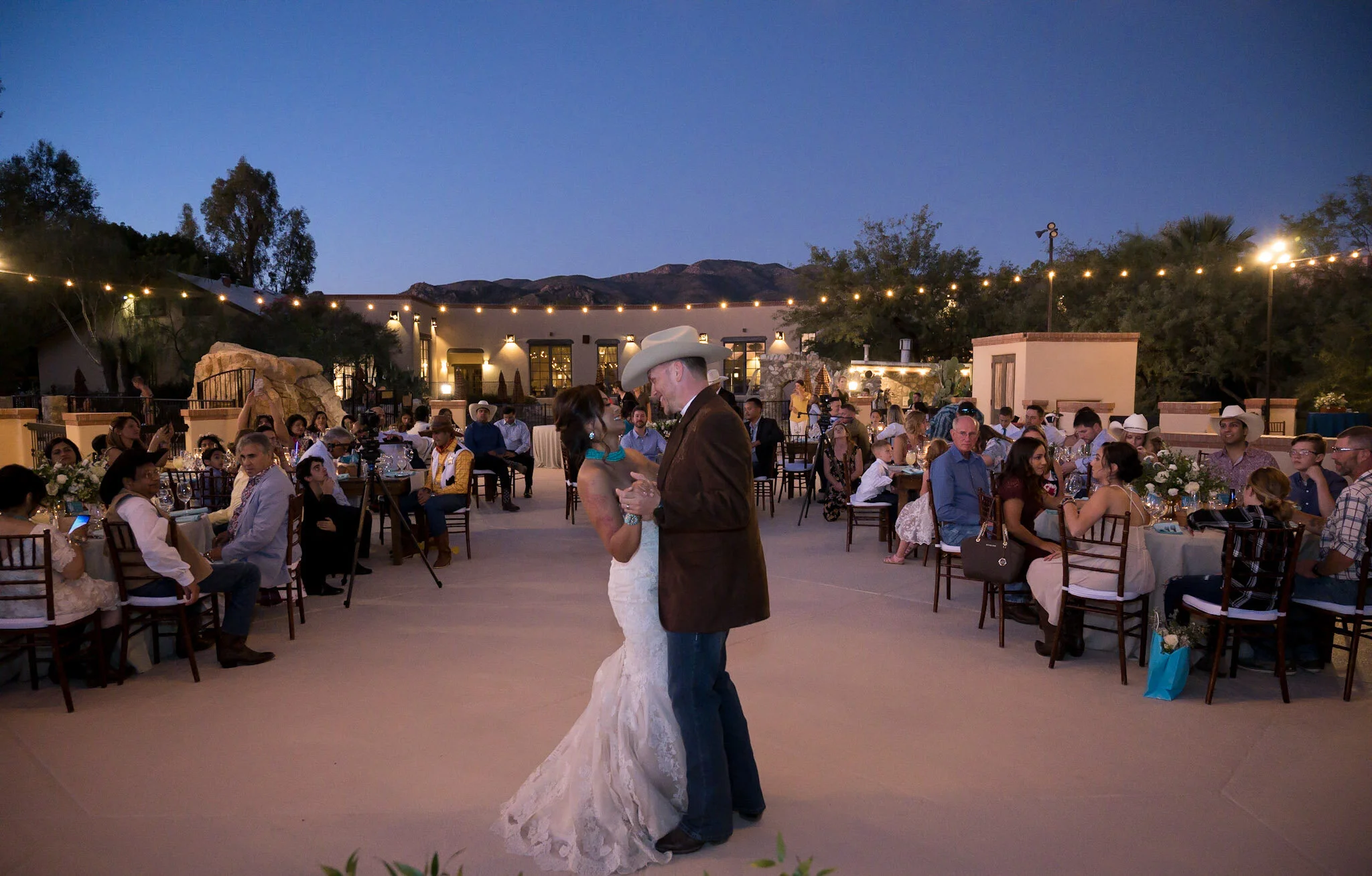 A bride and groom dancing at an outdoor wedding reception during evening, with guests seated at tables and string lights overhead. Mountains and trees are visible in the background.