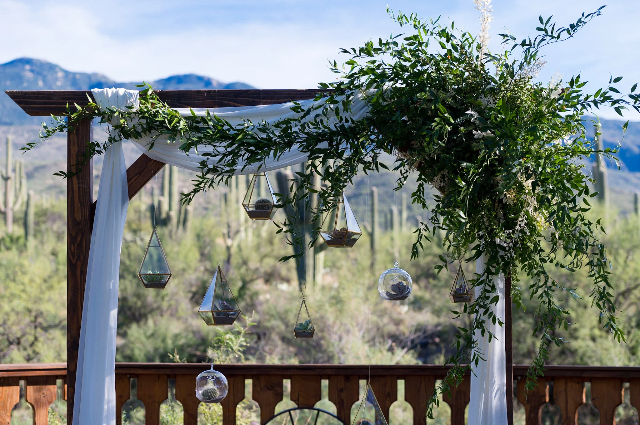 Outdoor pergola decorated with white fabric drapery, lush greenery, and hanging terrarium planters, with a mountain range in the background.
