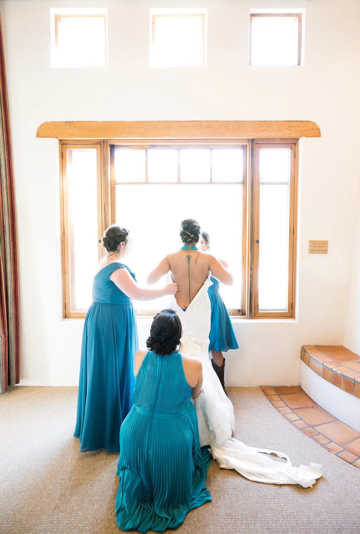 Bridesmaids helping bride put on wedding dress in front of a window with sunlight shining through.