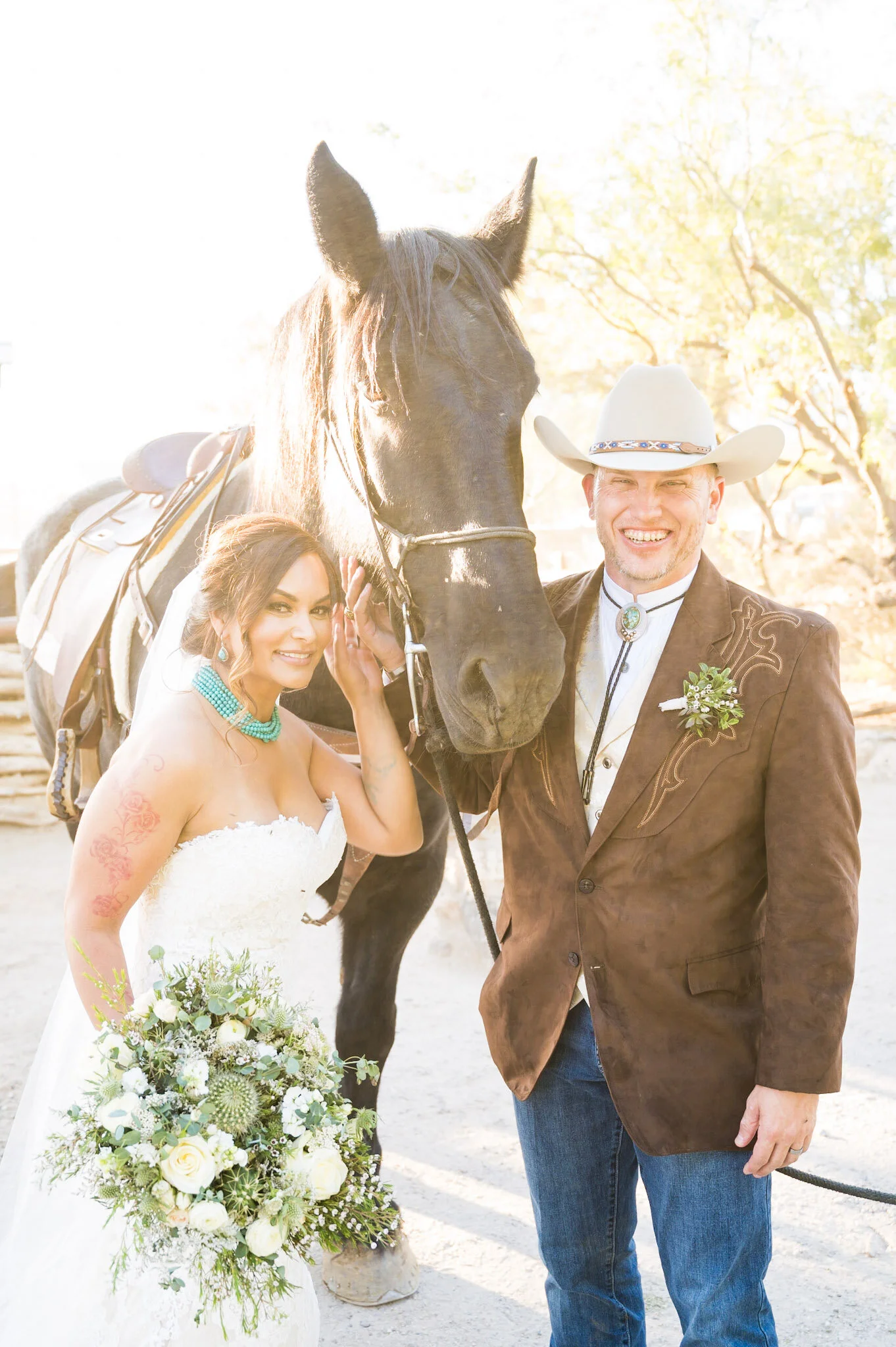 A wedding scene with a bride, groom, and a horse. The bride has dark hair, is wearing a white strapless wedding dress, and holding a bouquet of white and green flowers. The groom is dressed in a brown leather jacket, cowboy hat, and bolo tie, smiling