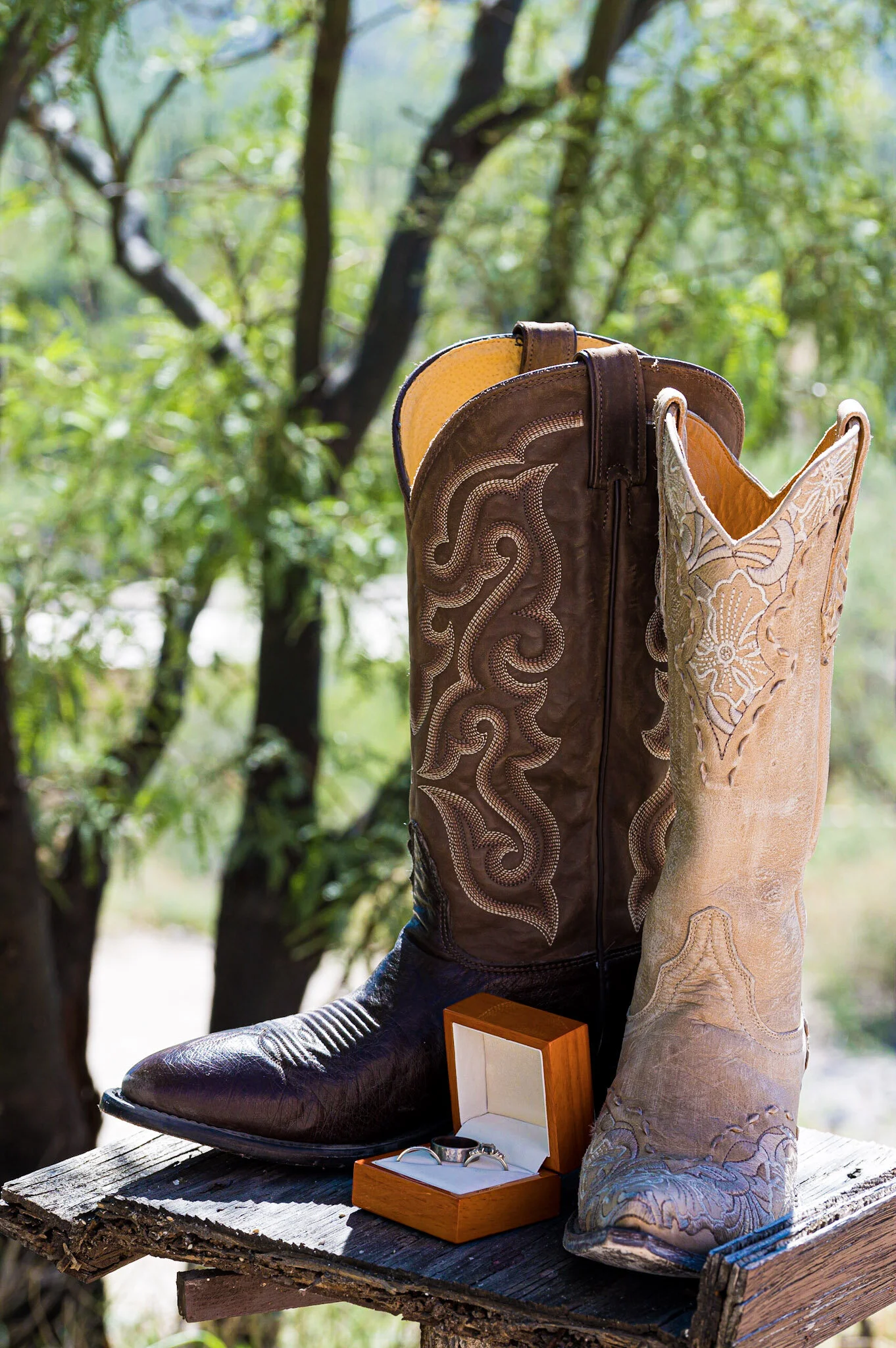 A cowboy boot, a hat, and a jewelry box with a ring, all placed on a wooden surface with a blurred green outdoor background.