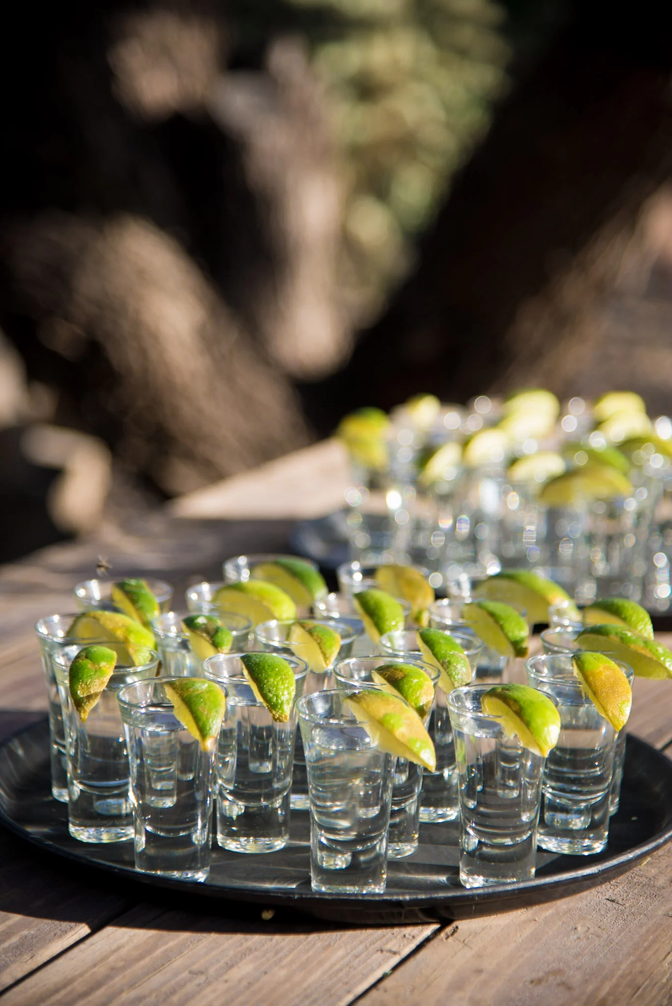 Small glasses filled with sliced lime wedges on a black tray, set on a wooden surface outdoors.
