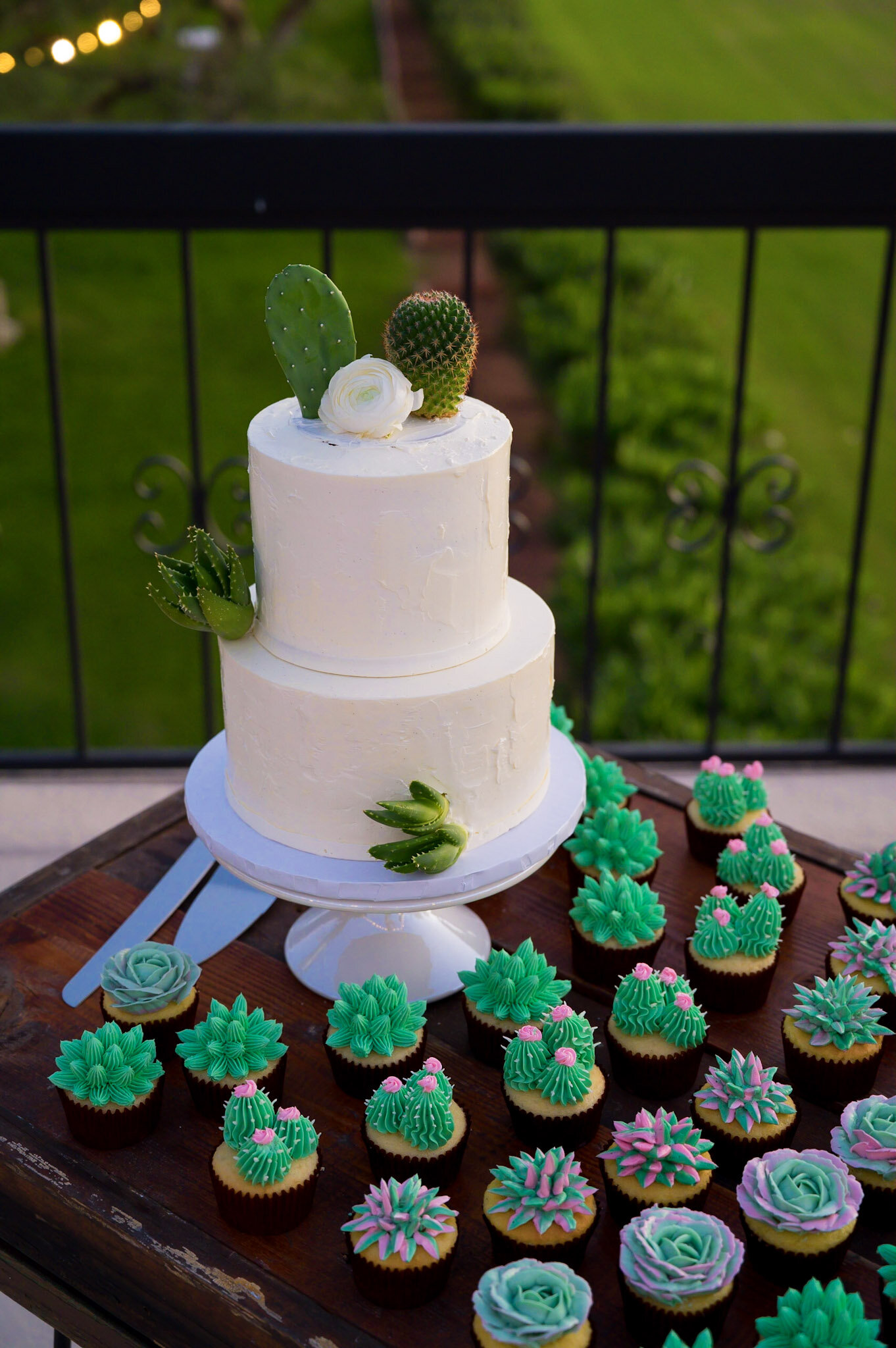A white, two-tiered cake decorated with cactus-shaped toppers, set on a white cake stand, surrounded by blue and pink decorated cupcakes with various floral and cactus-inspired frosting designs on a wooden table outdoors.