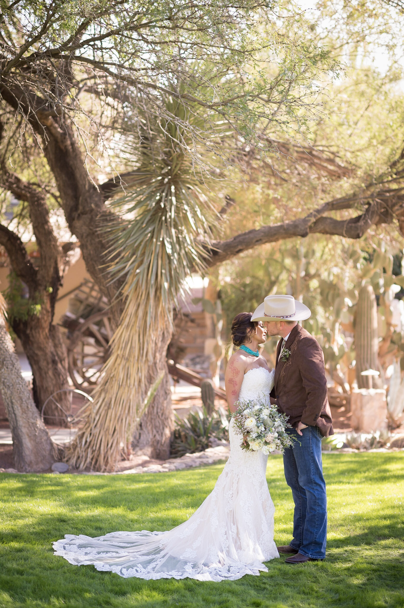 A bride and groom standing close together in a garden, surrounded by trees and greenery, with the bride holding a bouquet of flowers and wearing a white lace wedding dress, and the groom wearing a cowboy hat, brown blazer, and jeans.