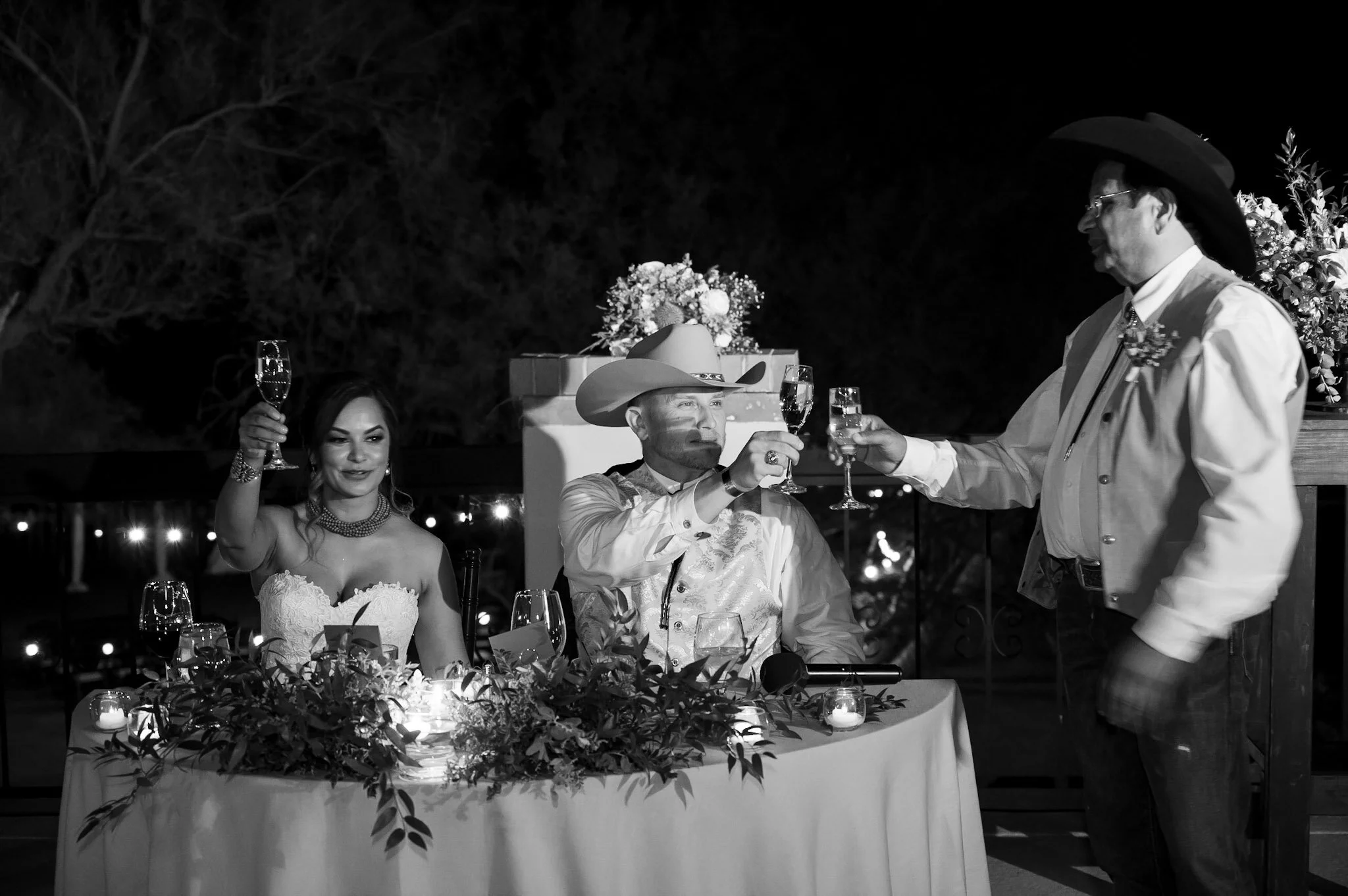 A black and white photo of a wedding reception at night. A bride and groom are seated at a decorated table with flowers and candles. The bride is holding a glass, and there is a man in cowboy attire with a hat, vest, and floral boutonnière standing a
