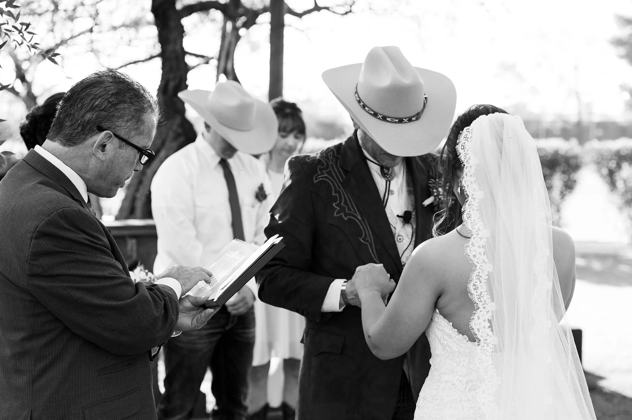 A black and white photo of a couple getting married outdoors. The bride, wearing a lace wedding dress and veil, and the groom, wearing a cowboy hat and western-style clothing, hold hands and look at each other. An officiant, reading from a book, stan