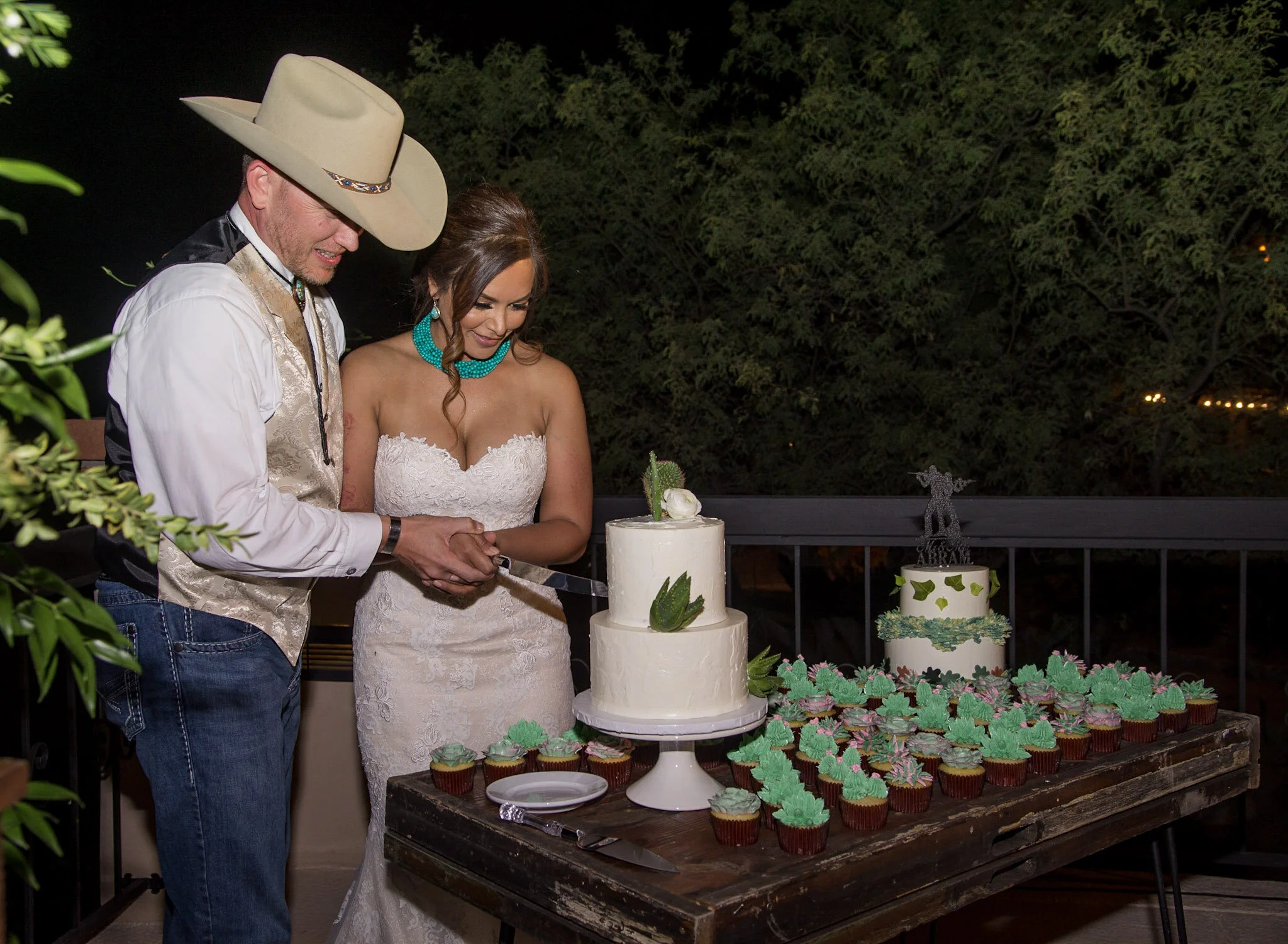 A man and woman, likely a bride and groom, cutting a wedding cake at an outdoor reception at night. The man is wearing a large beige cowboy hat, and the woman is dressed in a white lace wedding gown and turquoise jewelry. The table has a multi-layere