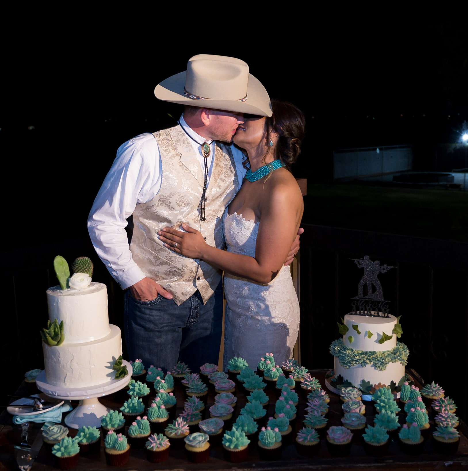 A couple wearing wedding attire kissing at their outdoor wedding celebration. The groom wears a cowboy hat and a paisley vest, while the bride wears a strapless white gown and turquoise jewelry. They stand behind two white wedding cakes decorated wit