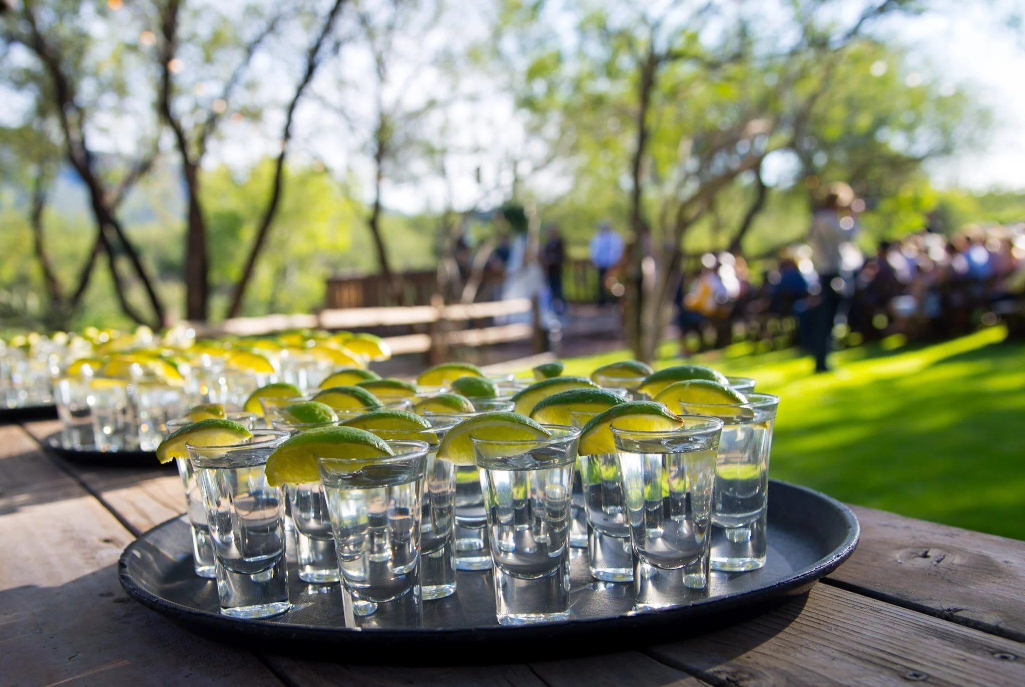 Tray of small glasses filled with clear liquid and garnished with lemon slices, set outdoors with blurred trees and people in the background.