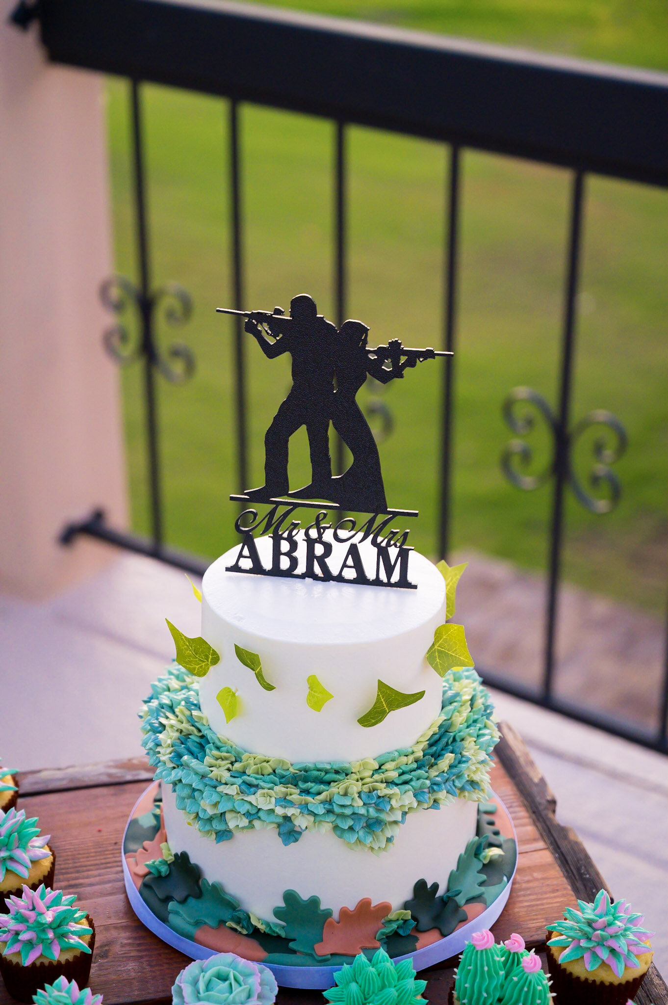 A wedding cake with a black topper of two soldiers holding rifles, with the words 'Mr & Mrs ABRAM' underneath. The cake is decorated with green and blue icing, and is surrounded by colorful cupcakes with various piping designs.