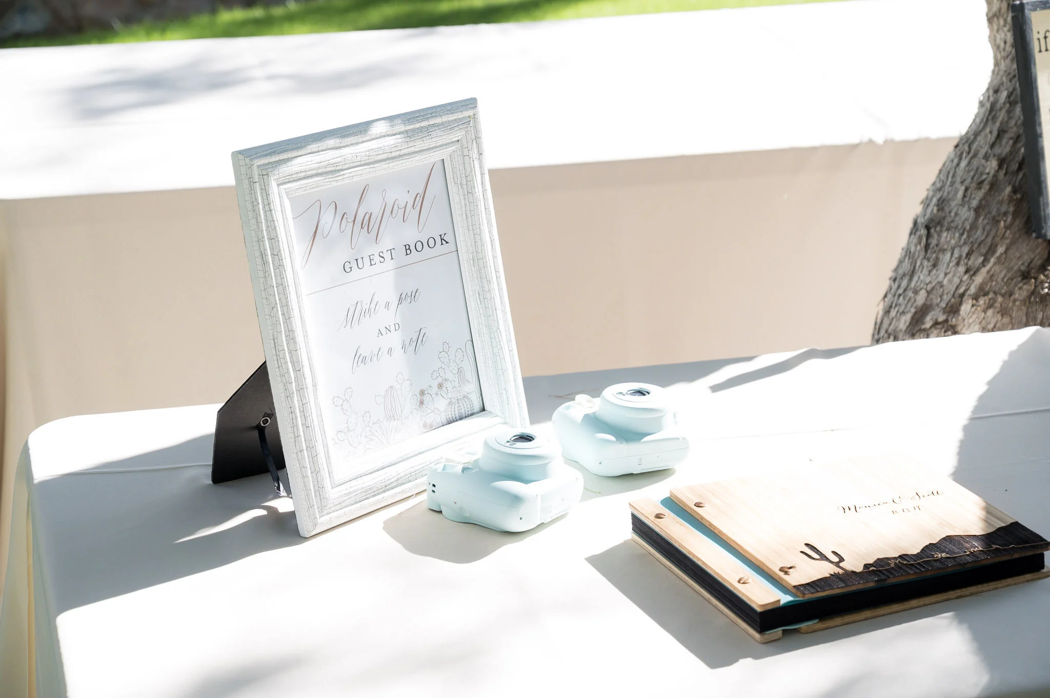 A guest book table at an outdoor event features a white framed sign labeled 'Polaroid Guest Book' with handwritten instructions to take a photo and leave a note, flanked by white decorative cameras and a wooden sign, with a tree and grass in the back